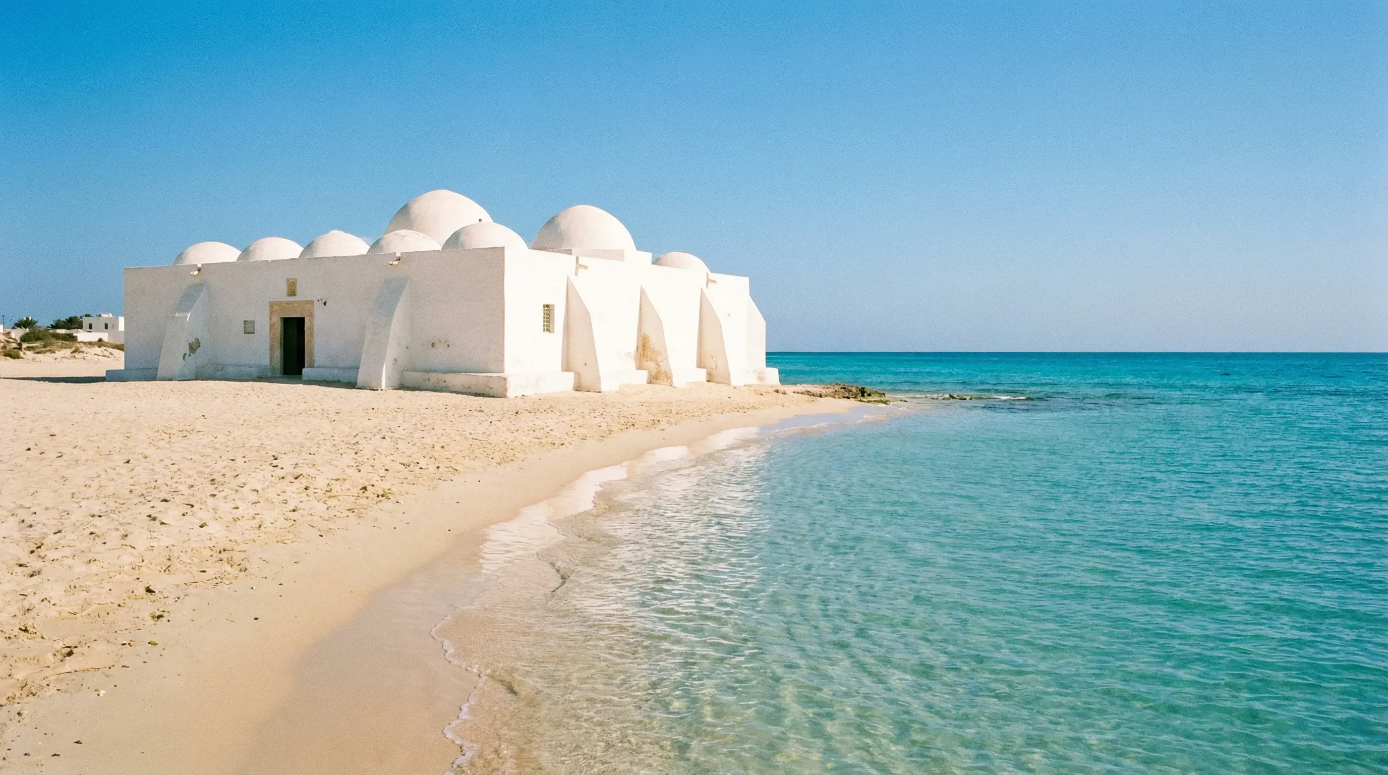 A white domed marabout building on a sandy beach next to turquoise water under a clear sky in Djerba, Tunisia.