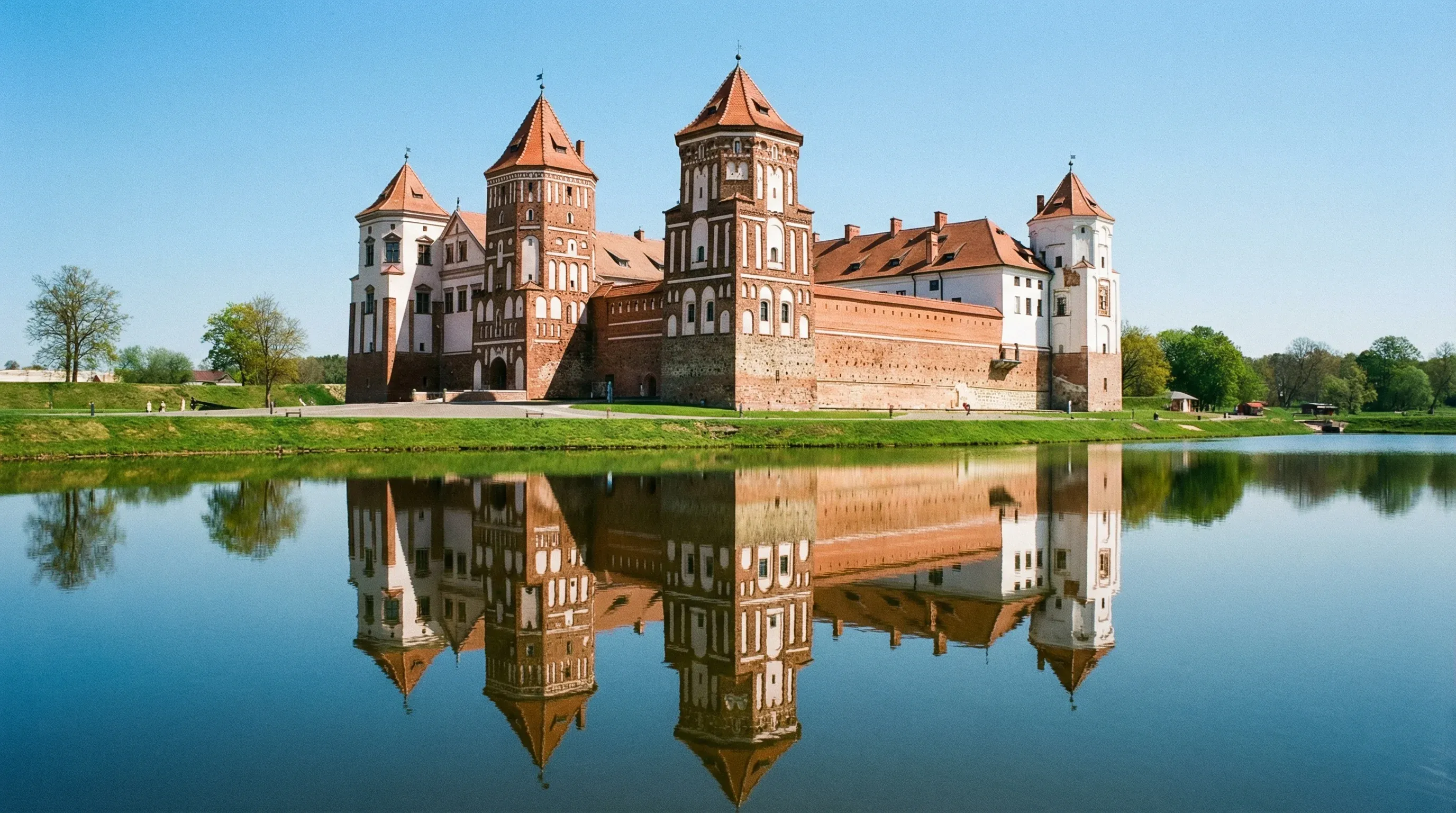 The red brick and white stone towers of Mir Castle reflected in a calm pond under bright sunlight.