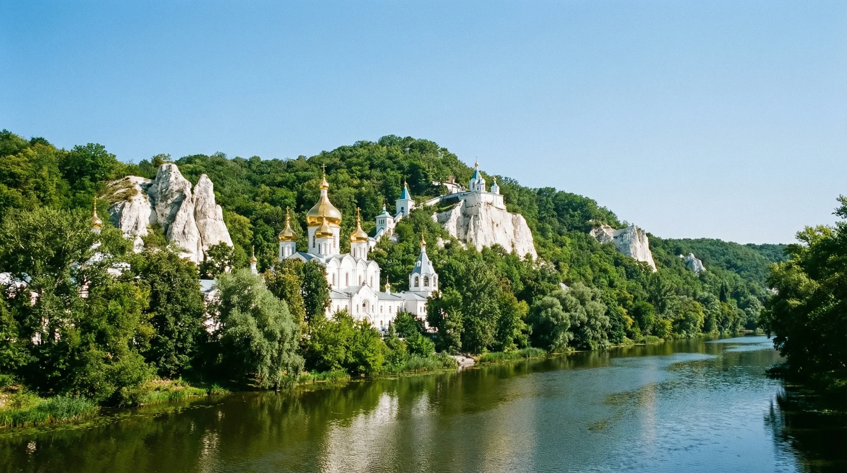 The white monastery buildings and golden domes of the Holy Mountains Lavra built into chalk cliffs above a river.