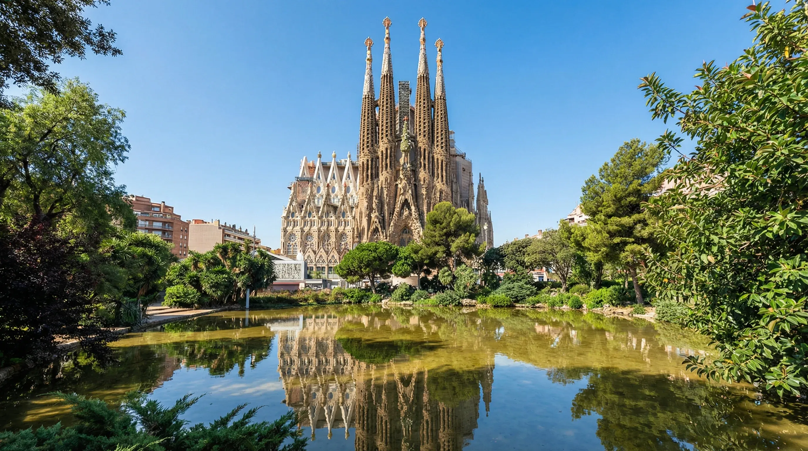 The Nativity facade of the Sagrada Familia basilica in Barcelona seen across a park pond under a clear sky.