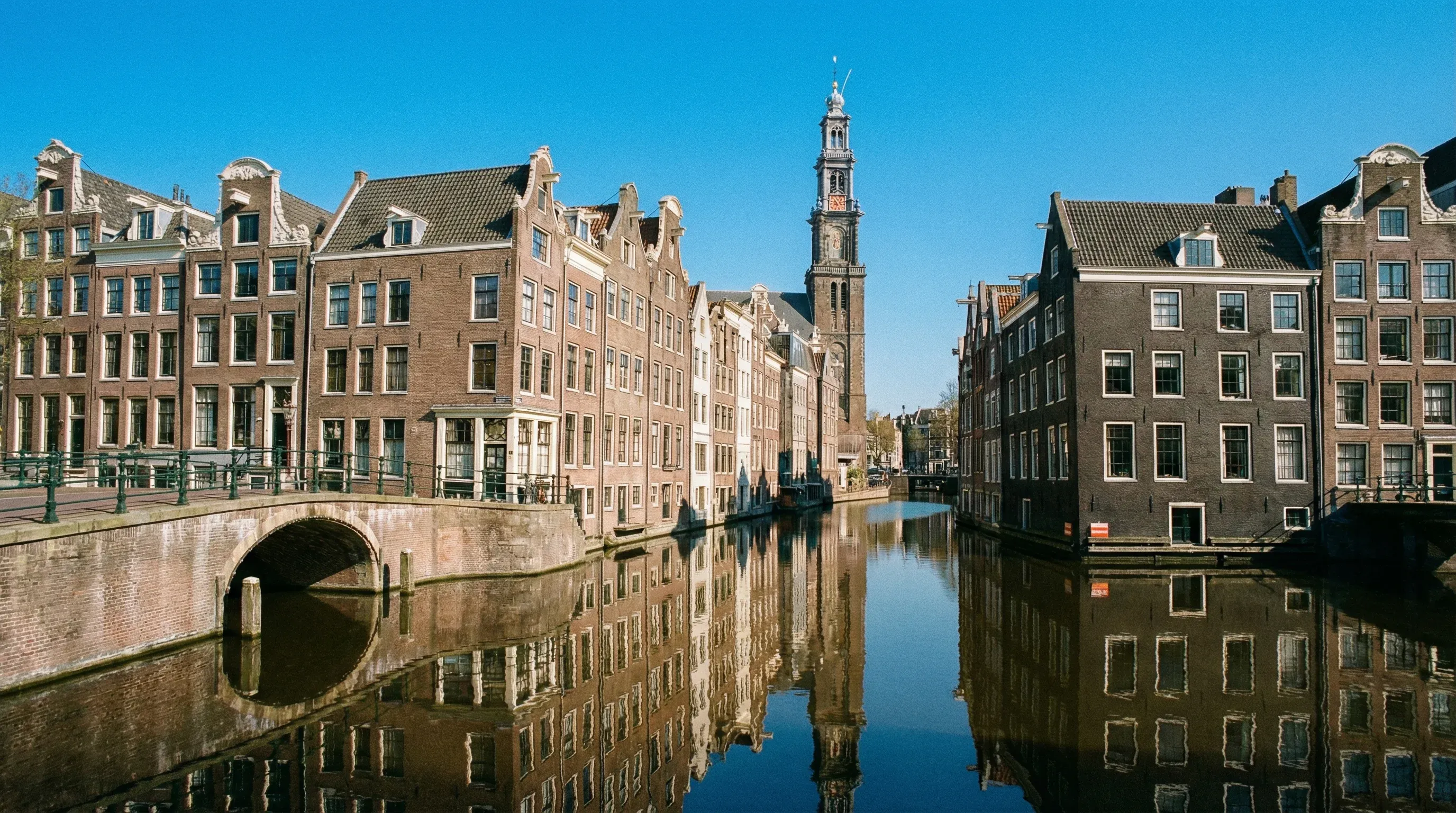 A view of the Prinsengracht canal in Amsterdam with historic gabled houses and the Westerkerk tower under a clear blue sky.