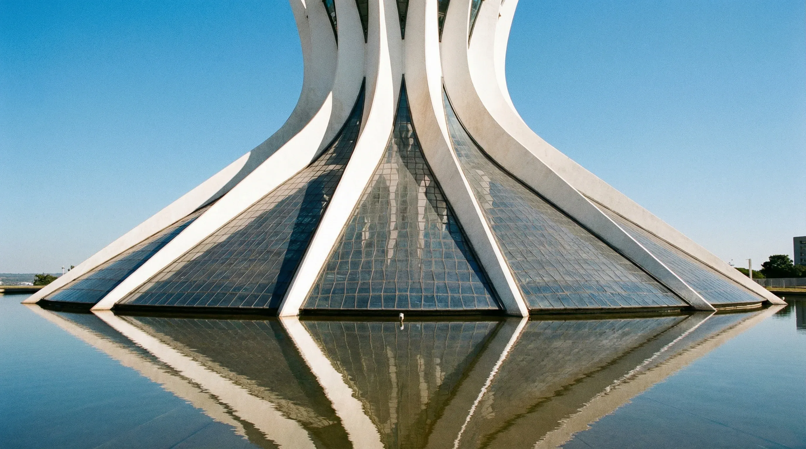 Exterior view of the modernist white concrete columns of the Cathedral of Brasília against a clear blue sky.