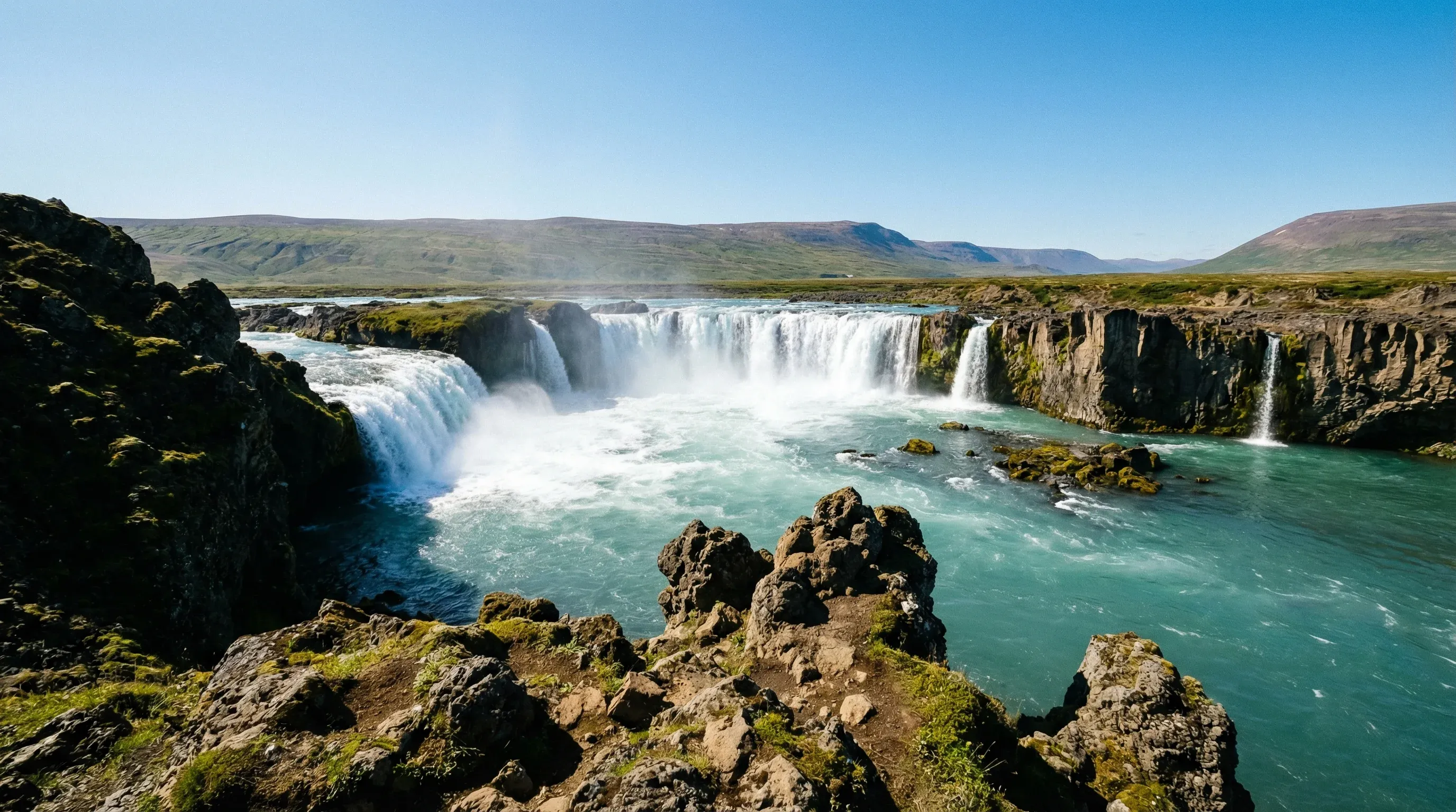 The horseshoe-shaped Goðafoss waterfall in North Iceland, showing white water falling into a river canyon under a clear sky.