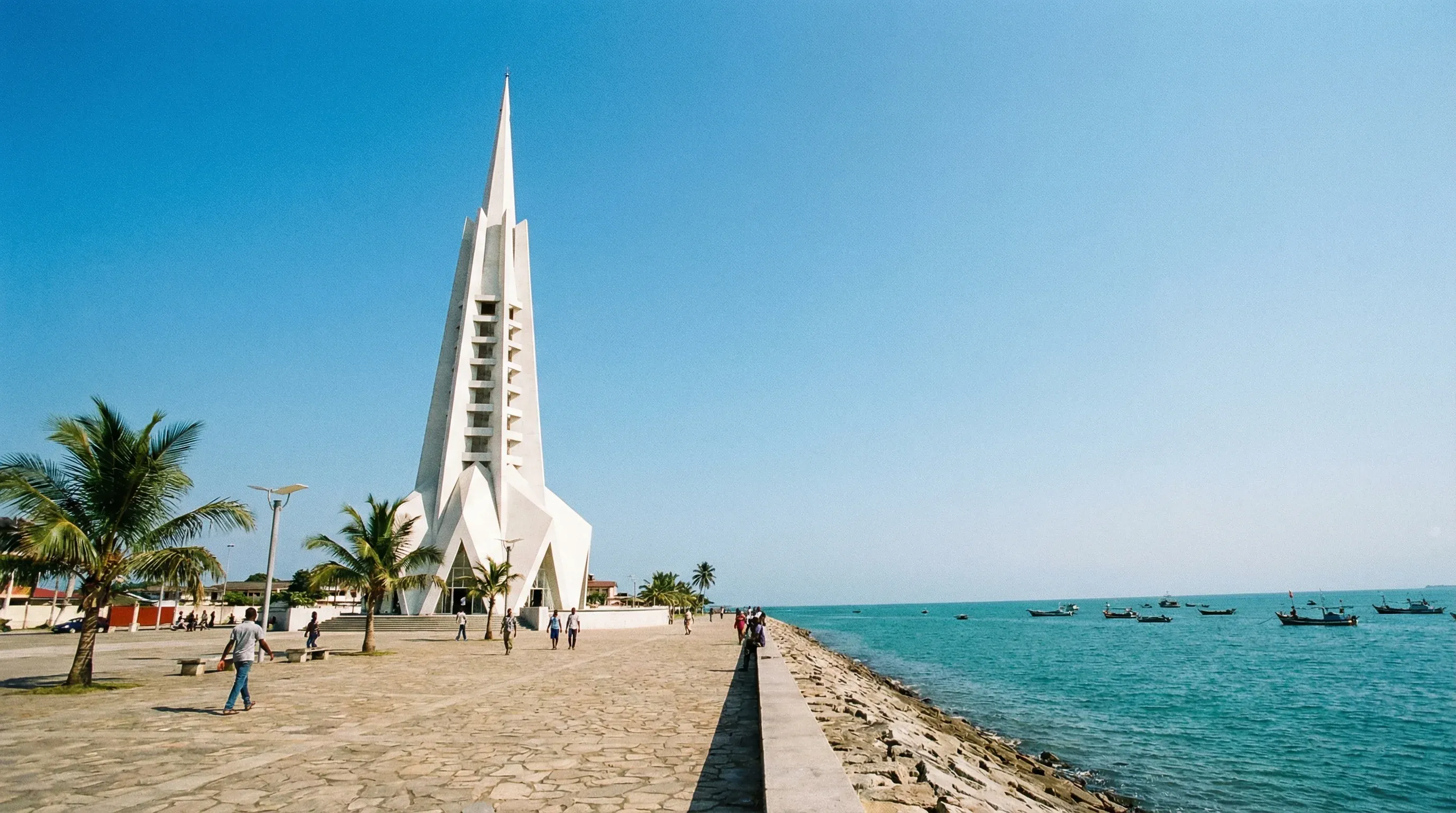 The tall, modern white Freedom Tower stands on the Bata waterfront promenade overlooking the Gulf of Guinea under a bright midday sky.