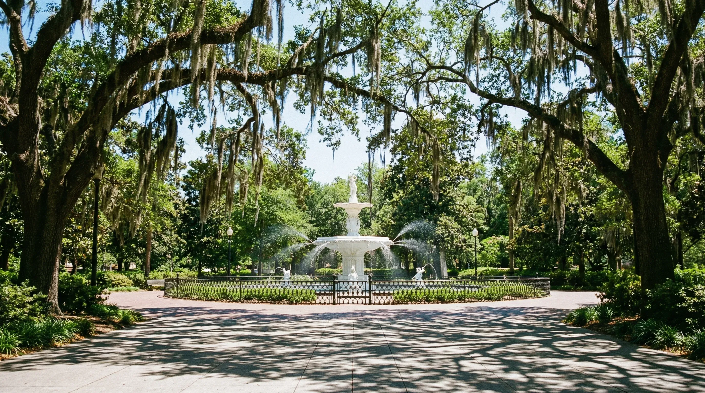 A large white ornamental fountain surrounded by oak trees with Spanish moss in a green park.