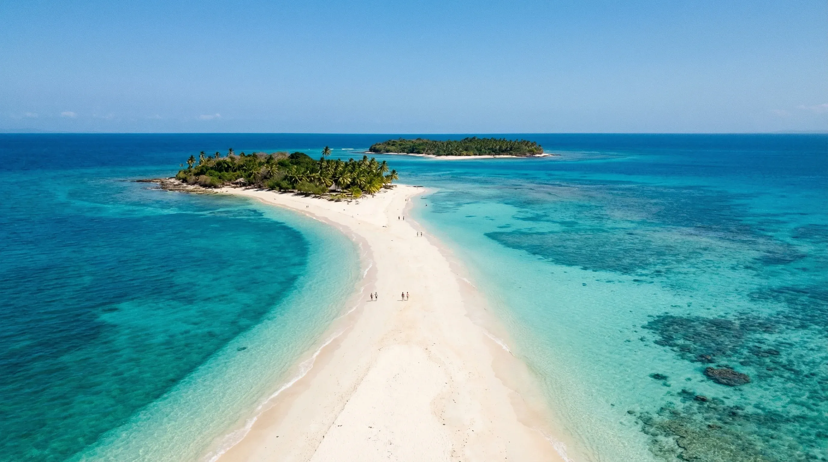 A high-angle photograph showing a long white sandbar connecting two tropical islands surrounded by turquoise water in Nosy Iranja.
