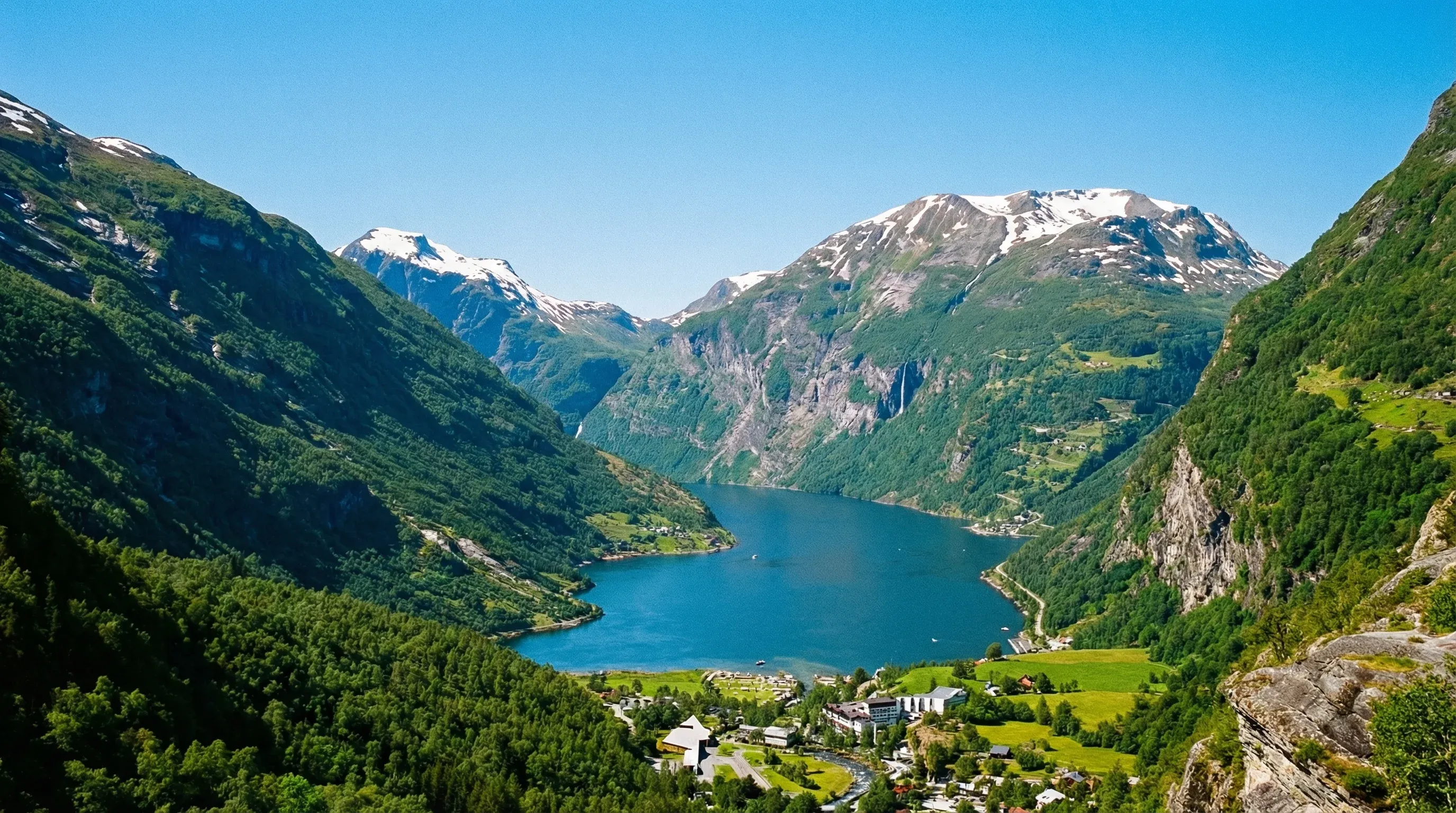 A wide-angle view of the deep blue Geirangerfjord surrounded by steep green mountains and waterfalls under a bright sun.