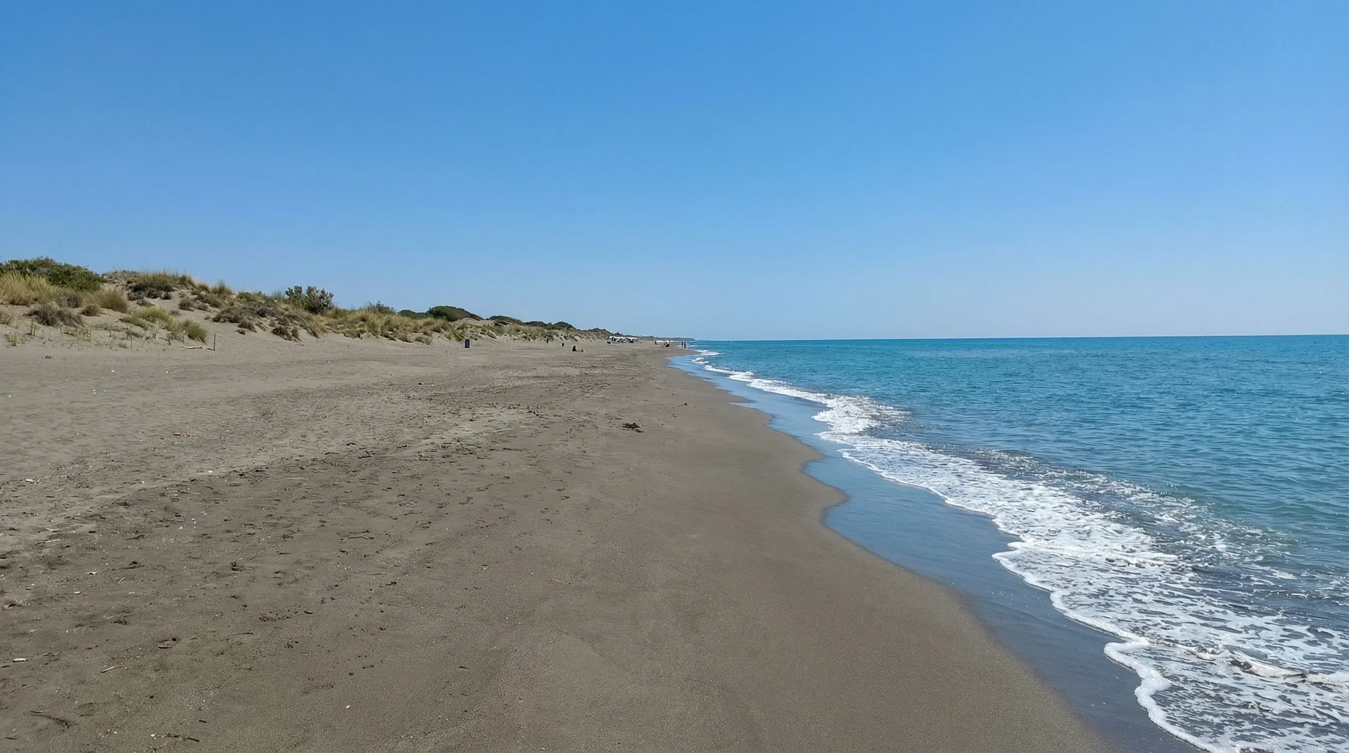 A wide, long stretch of dark sand beach meeting the blue Adriatic Sea under a clear sky near Ulcinj.