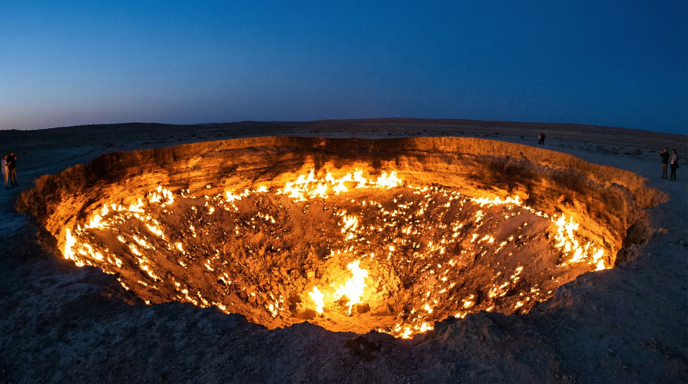 A large circular crater in the desert floor filled with hundreds of small orange gas fires burning at twilight.