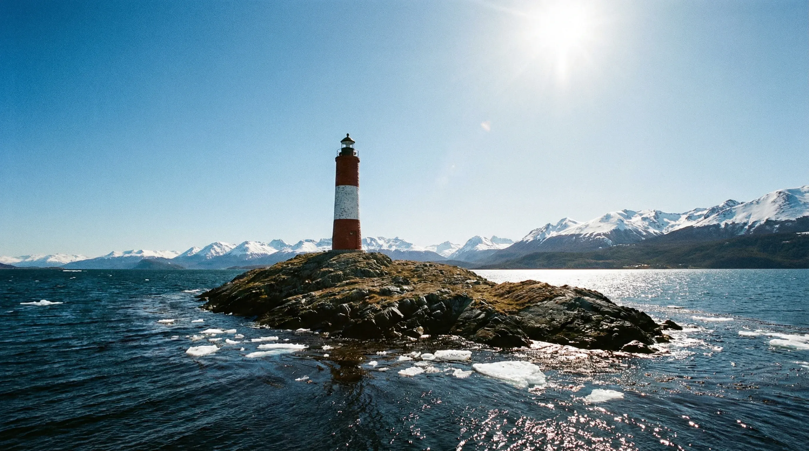 A red and white lighthouse stands on a rocky island in the Beagle Channel with mountains in the background.