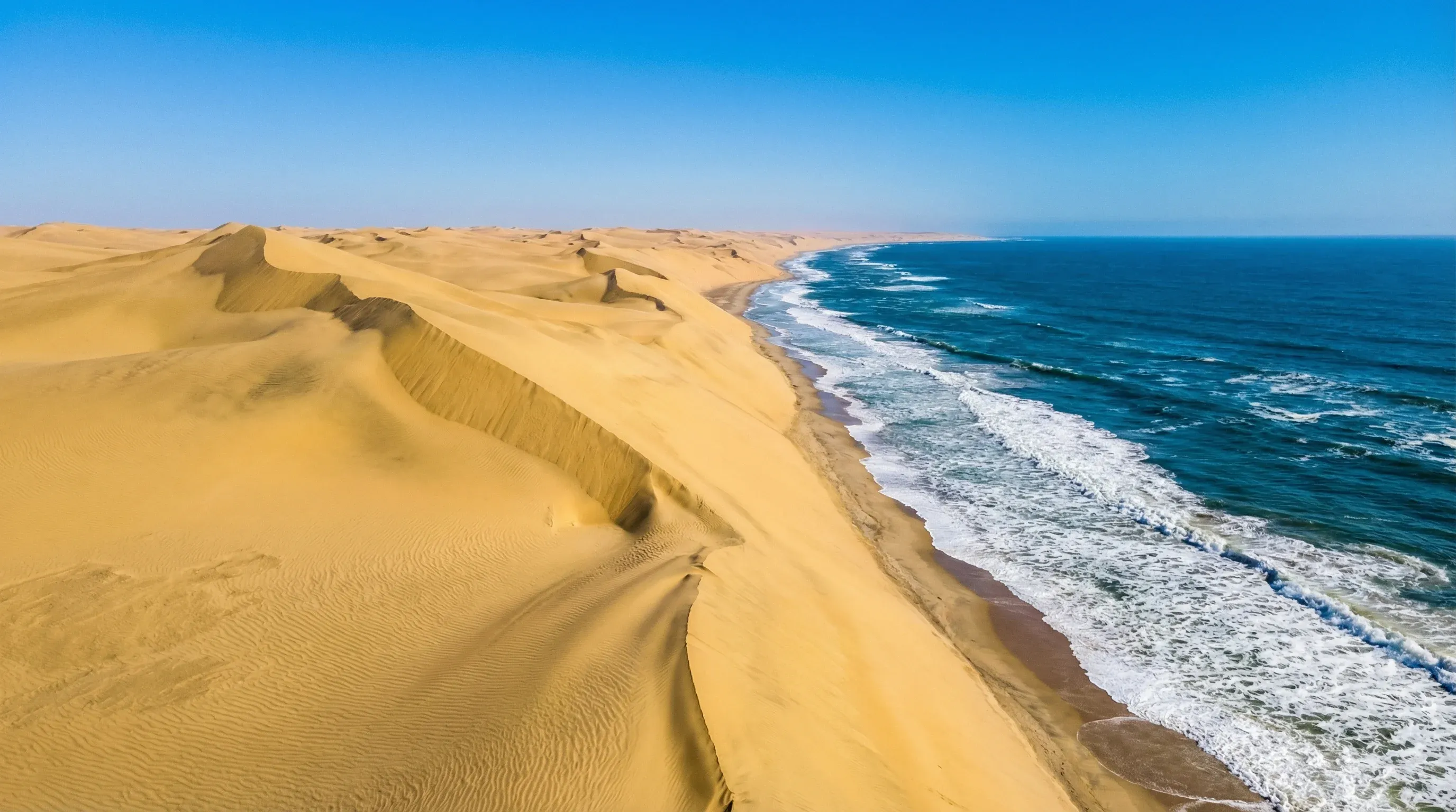 High yellow sand dunes of the Namib Desert meeting the blue Atlantic Ocean waves at Sandwich Harbour.