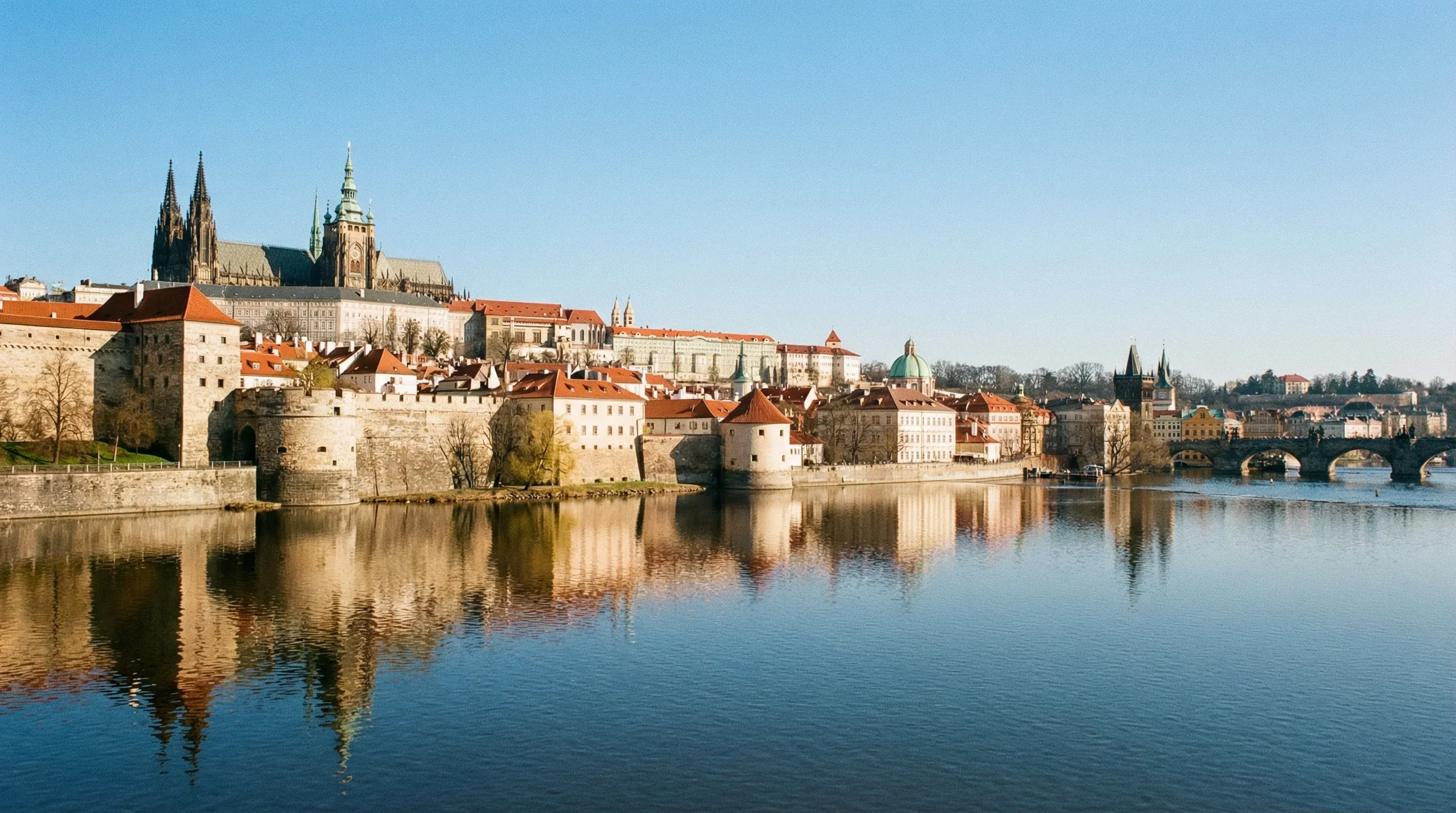 The historic complex of Prague Castle and the spires of St. Vitus Cathedral overlooking the Vltava River on a sunny day.
