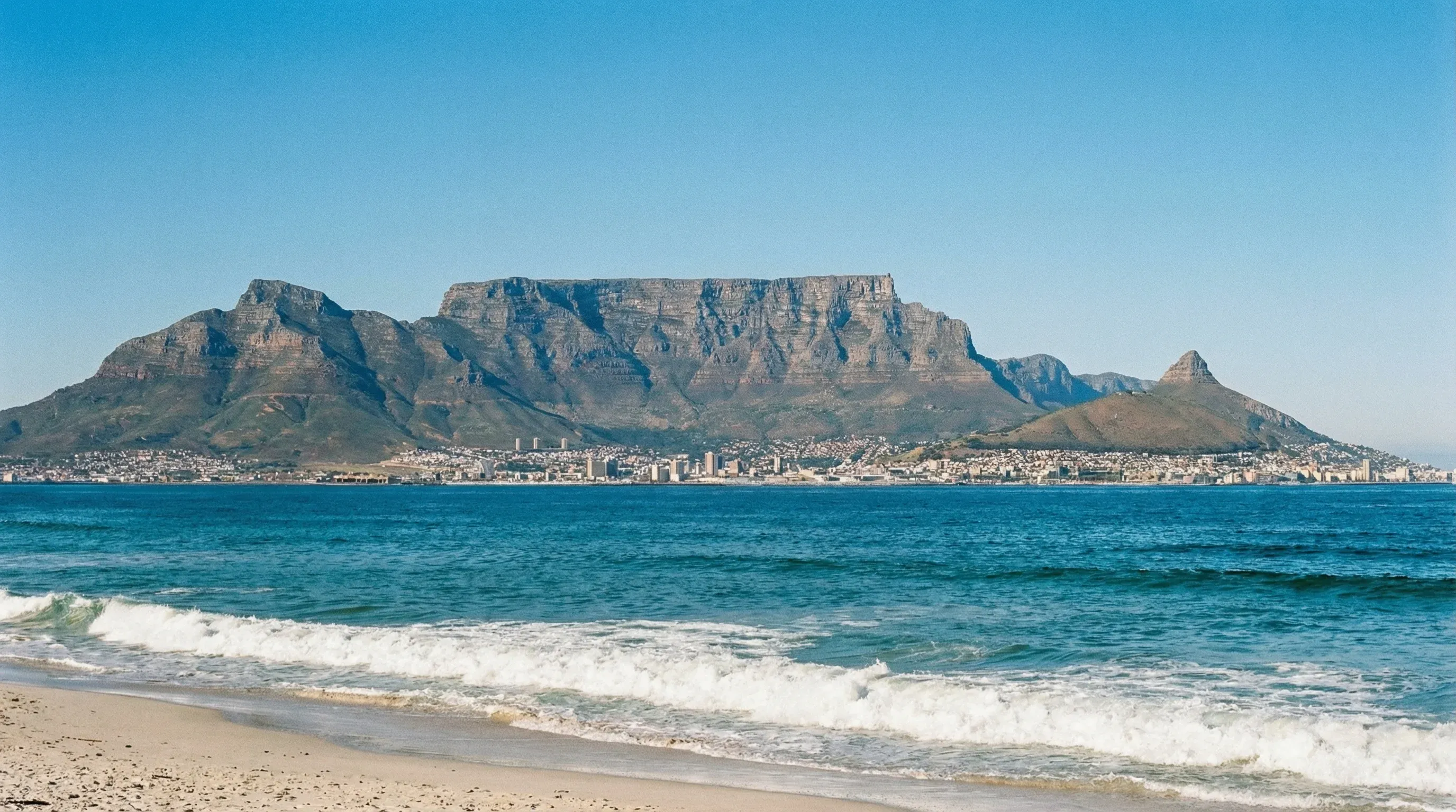 A view across the blue water of Table Bay towards the flat-topped Table Mountain and the city of Cape Town under a clear sky.