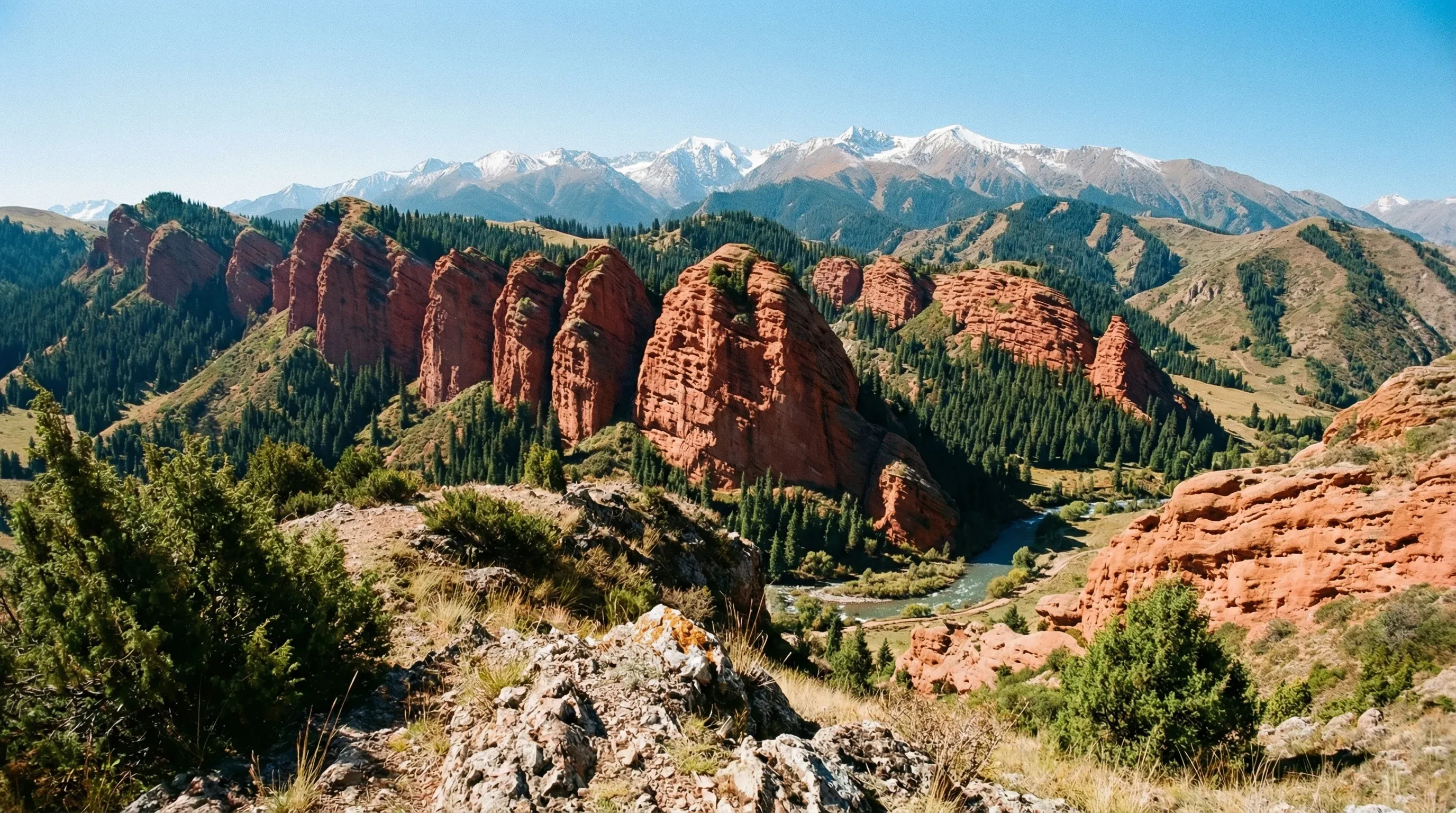 Red sandstone rock formations known as Jeti-Oguz with green pine trees and distant snow-covered mountains.