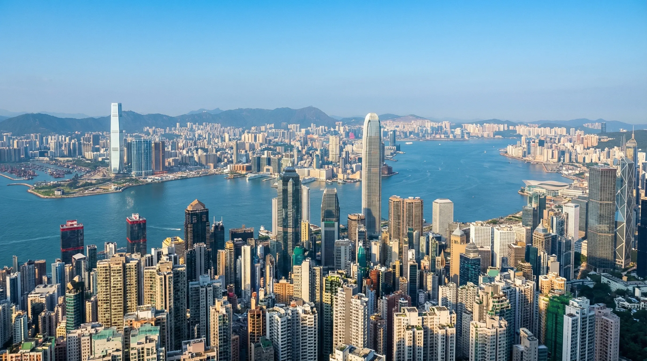 A high-angle view of the Hong Kong skyline and Victoria Harbour with skyscrapers and blue water.