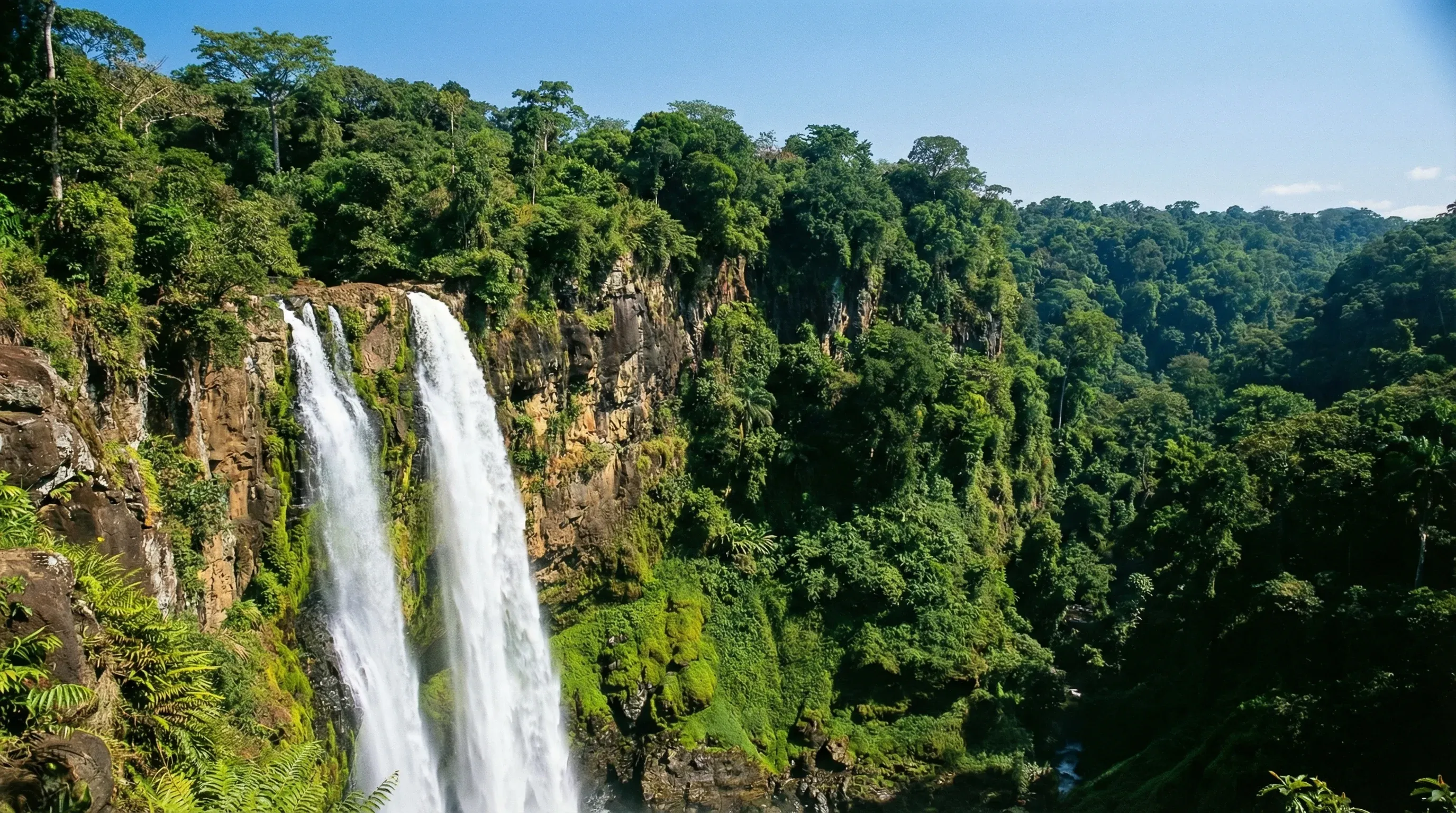 A wide-angle view of the massive twin Ekom-Nkam waterfalls cascading into a lush green rainforest basin.
