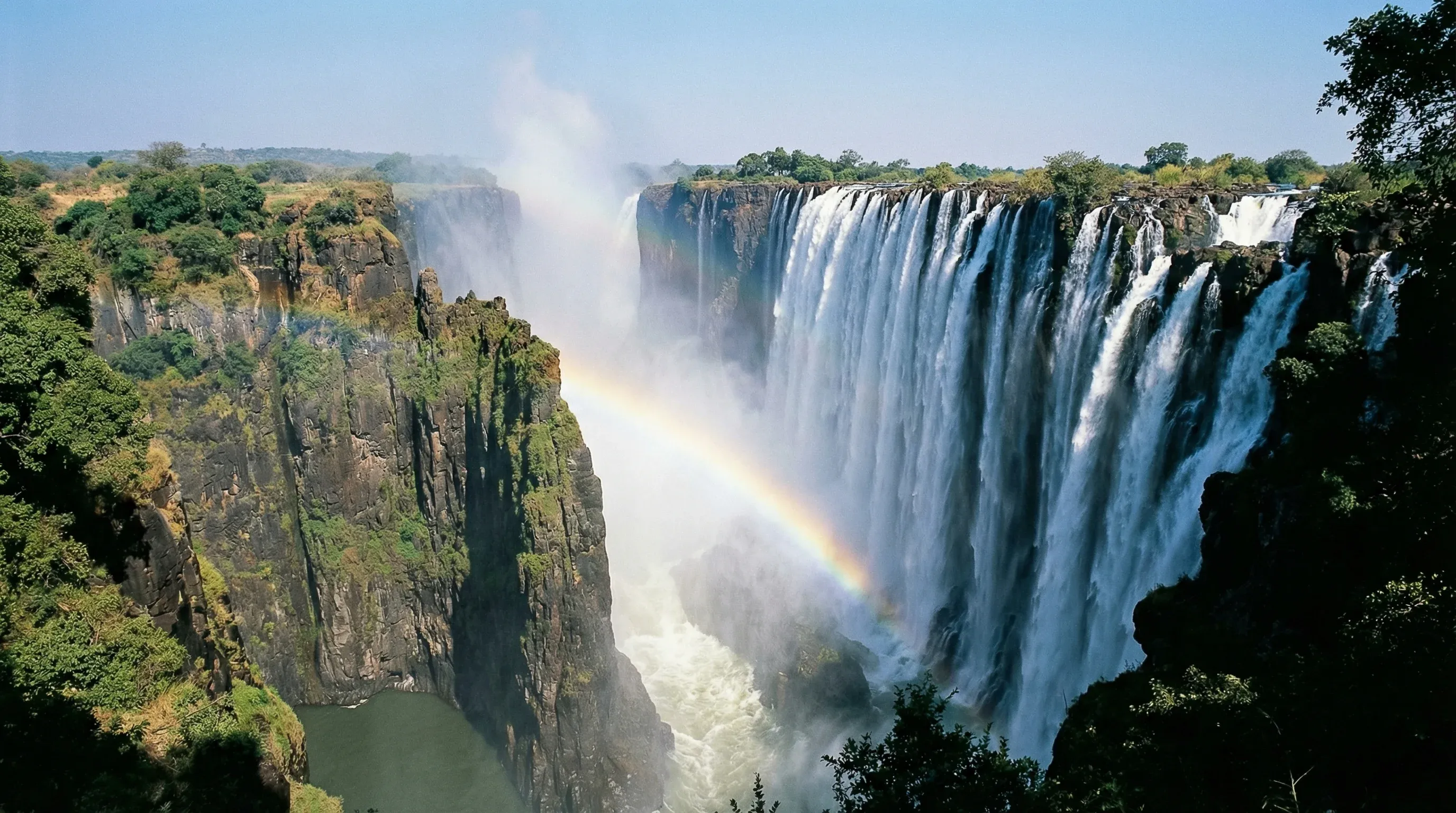 A wide-angle view of the Eastern Cataract of Victoria Falls in Zambia, showing the massive waterfall plunging into the Batoka Gorge under a clear sky.
