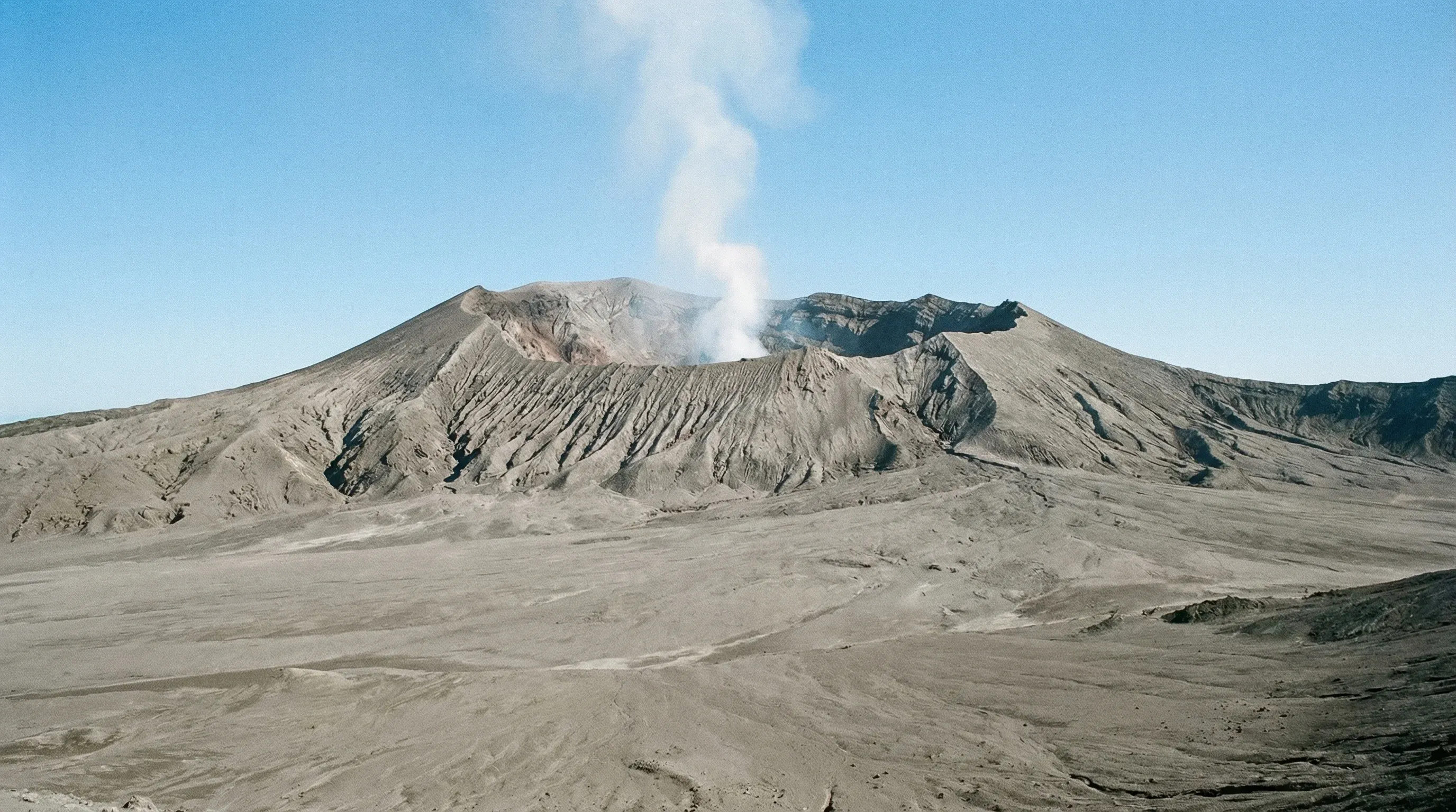 The grey volcanic cone of Mount Yasur rising from an ash desert under a bright blue sky on Tanna Island.