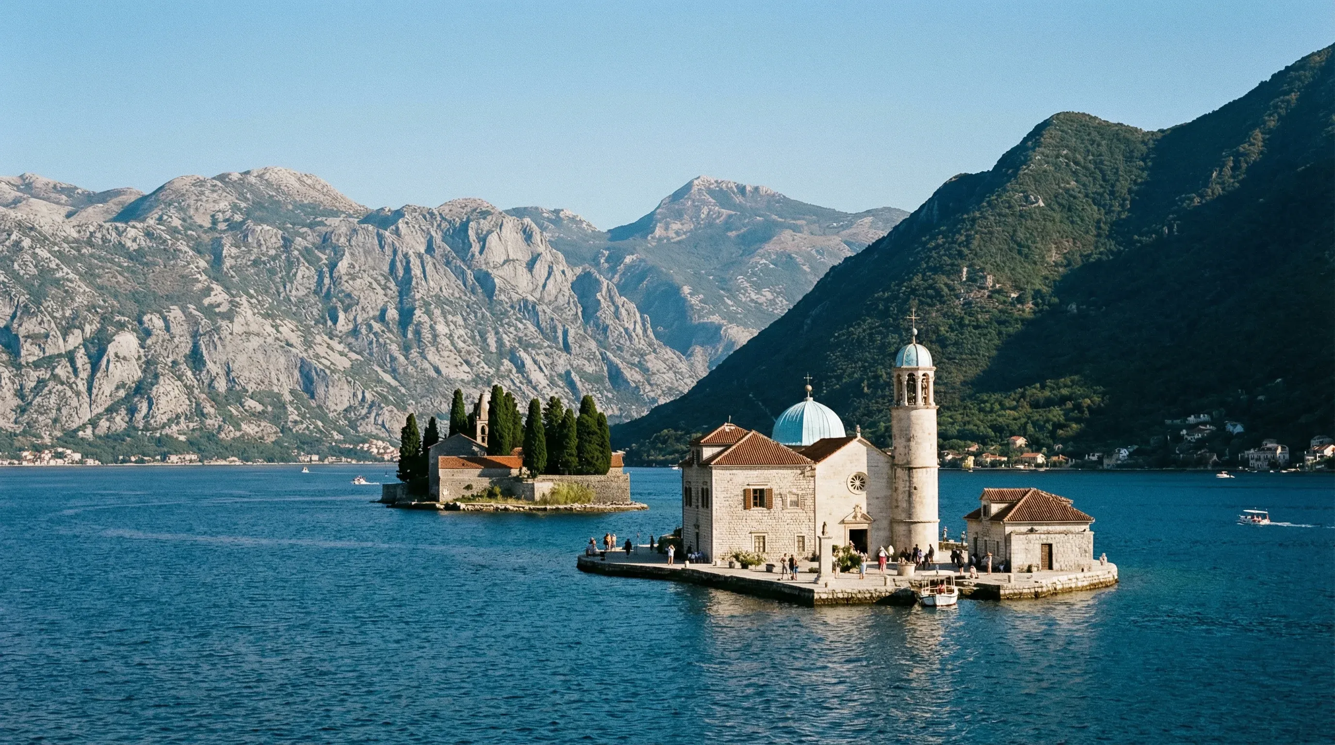A stone church with a blue dome on a small artificial island in the Bay of Kotor, surrounded by deep blue water and steep limestone mountains.