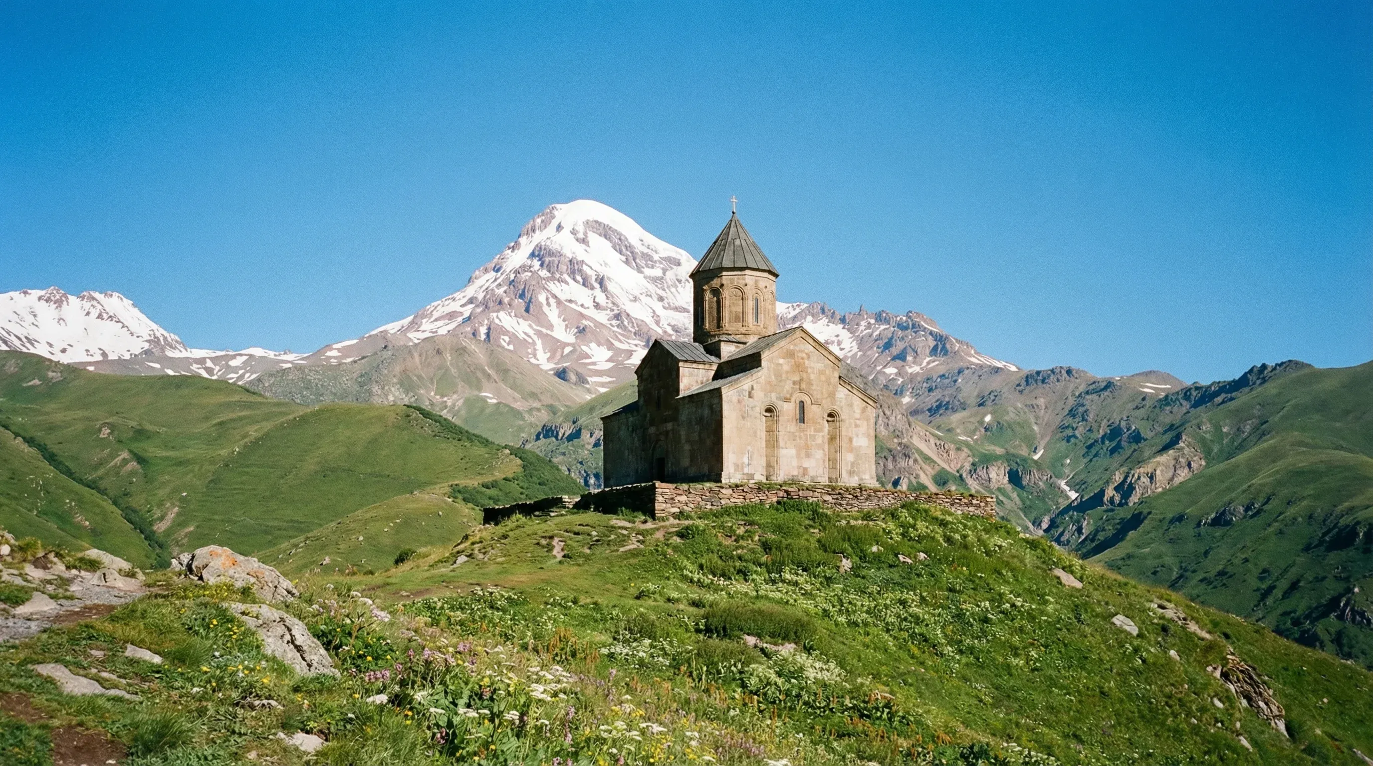 The stone Gergeti Trinity Church on a green hill with the snow-capped peak of Mount Kazbek in the background.
