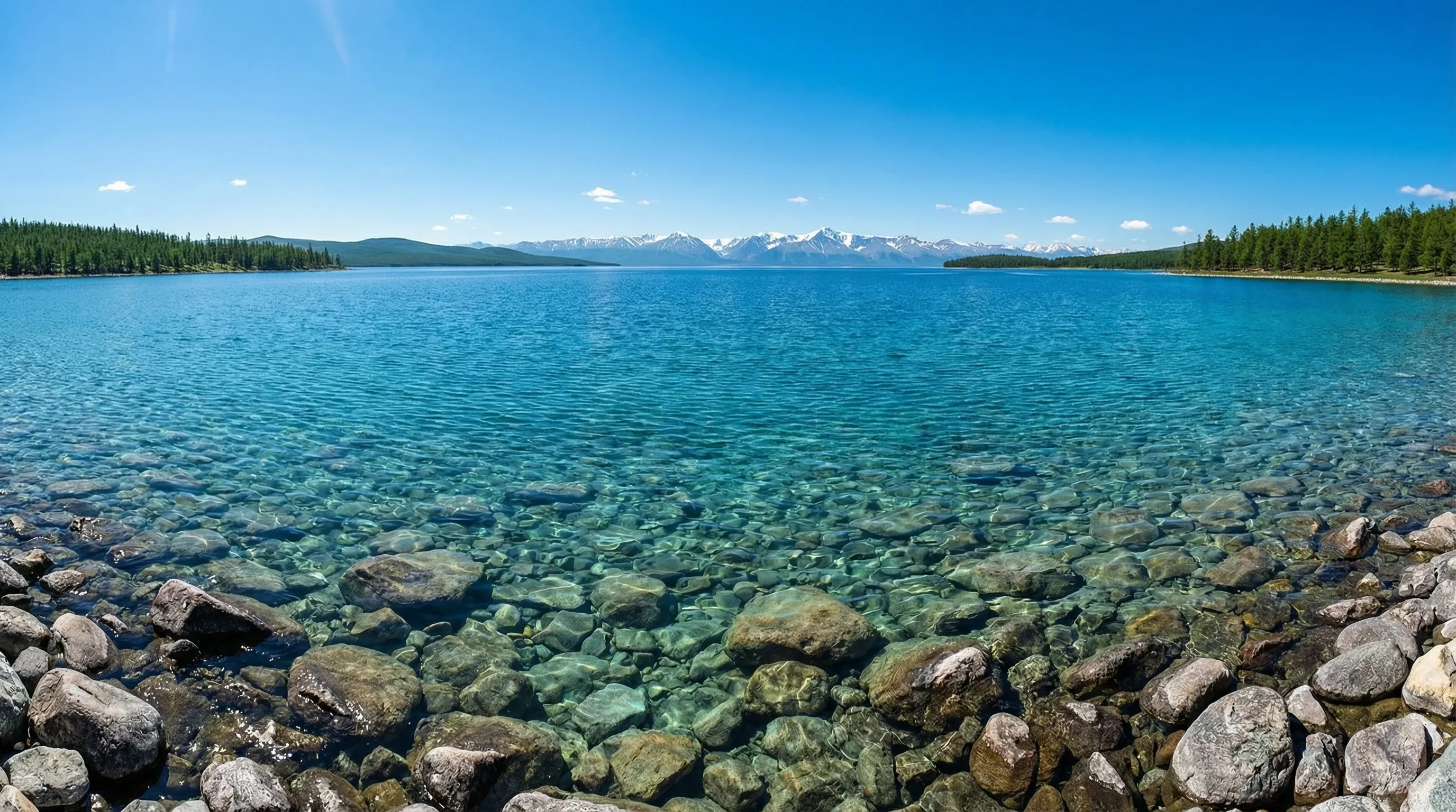 The clear blue waters of Lake Khövsgöl in Northern Mongolia, with a rocky shoreline in the foreground and mountains in the distance.