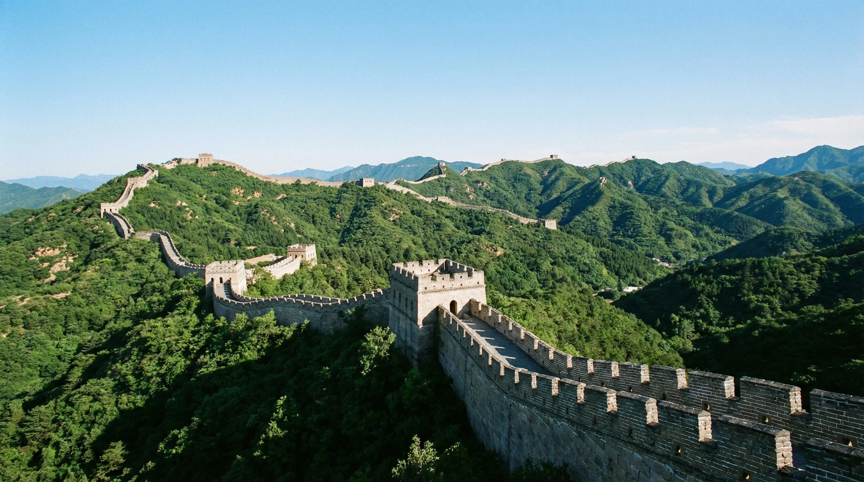 The stone Great Wall of China winding along the crest of green mountains under a bright blue sky.