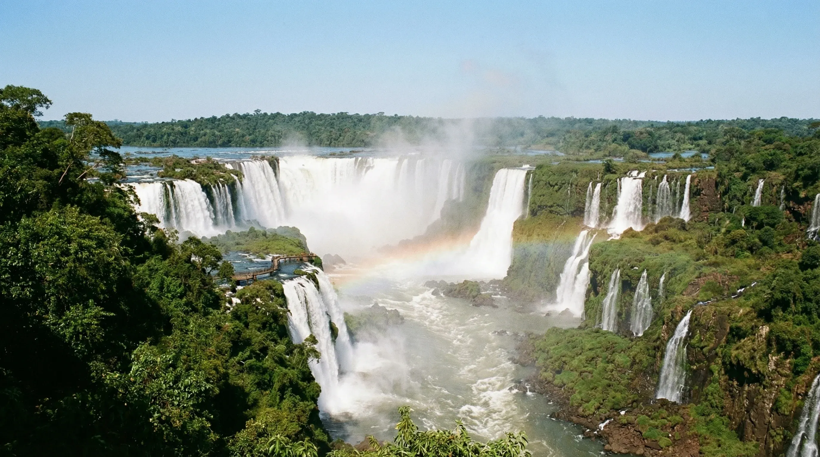 A wide view of the massive white waterfalls and green rainforest at Iguaçu Falls under a clear sky.