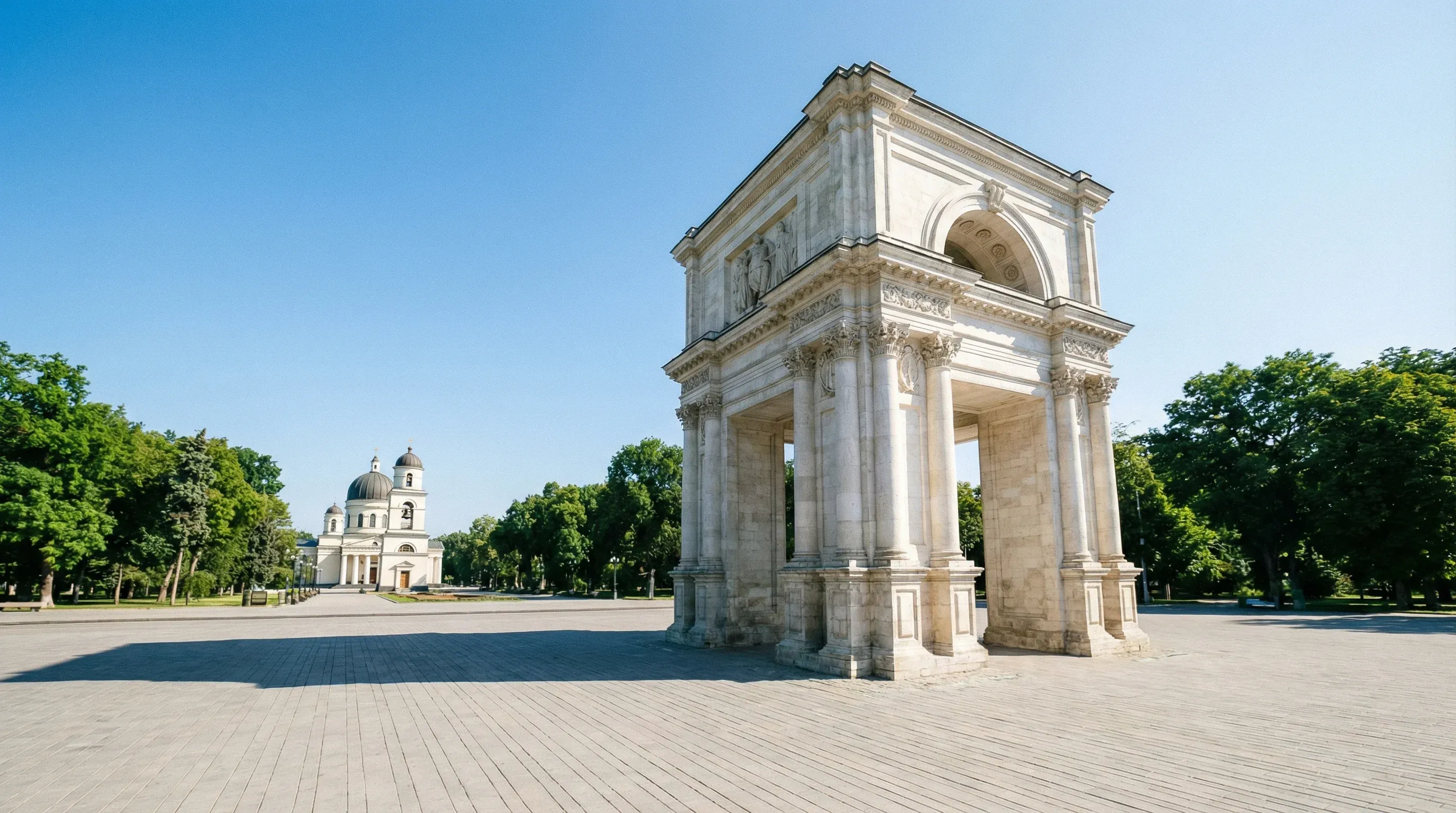 The white stone Triumphal Arch stands in a paved city square in Chișinău, with a cathedral dome visible in the background under a clear sky.