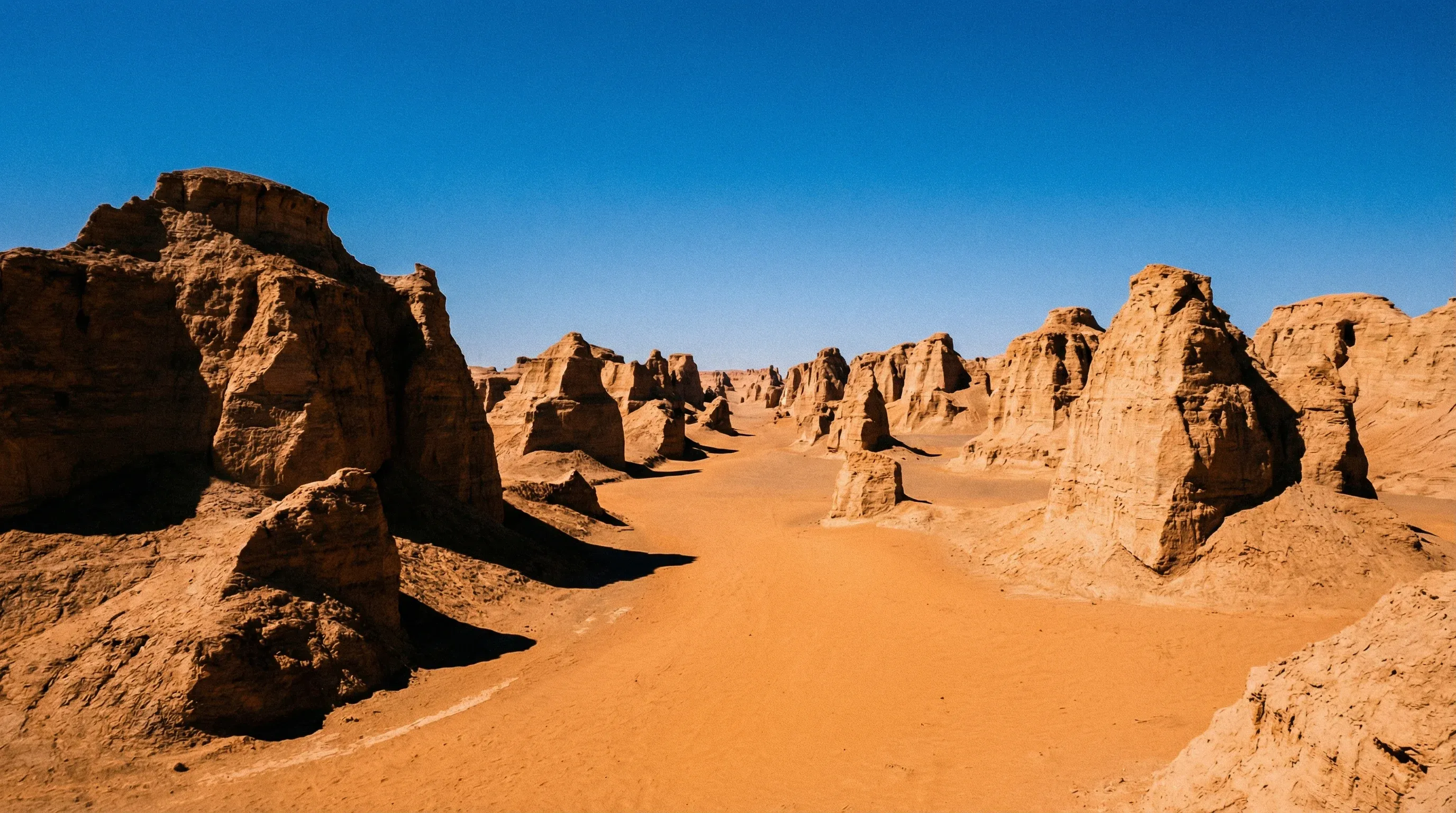 Large wind-eroded rock formations called yardangs rise from the sandy desert floor of the Lut Desert under a clear blue sky.