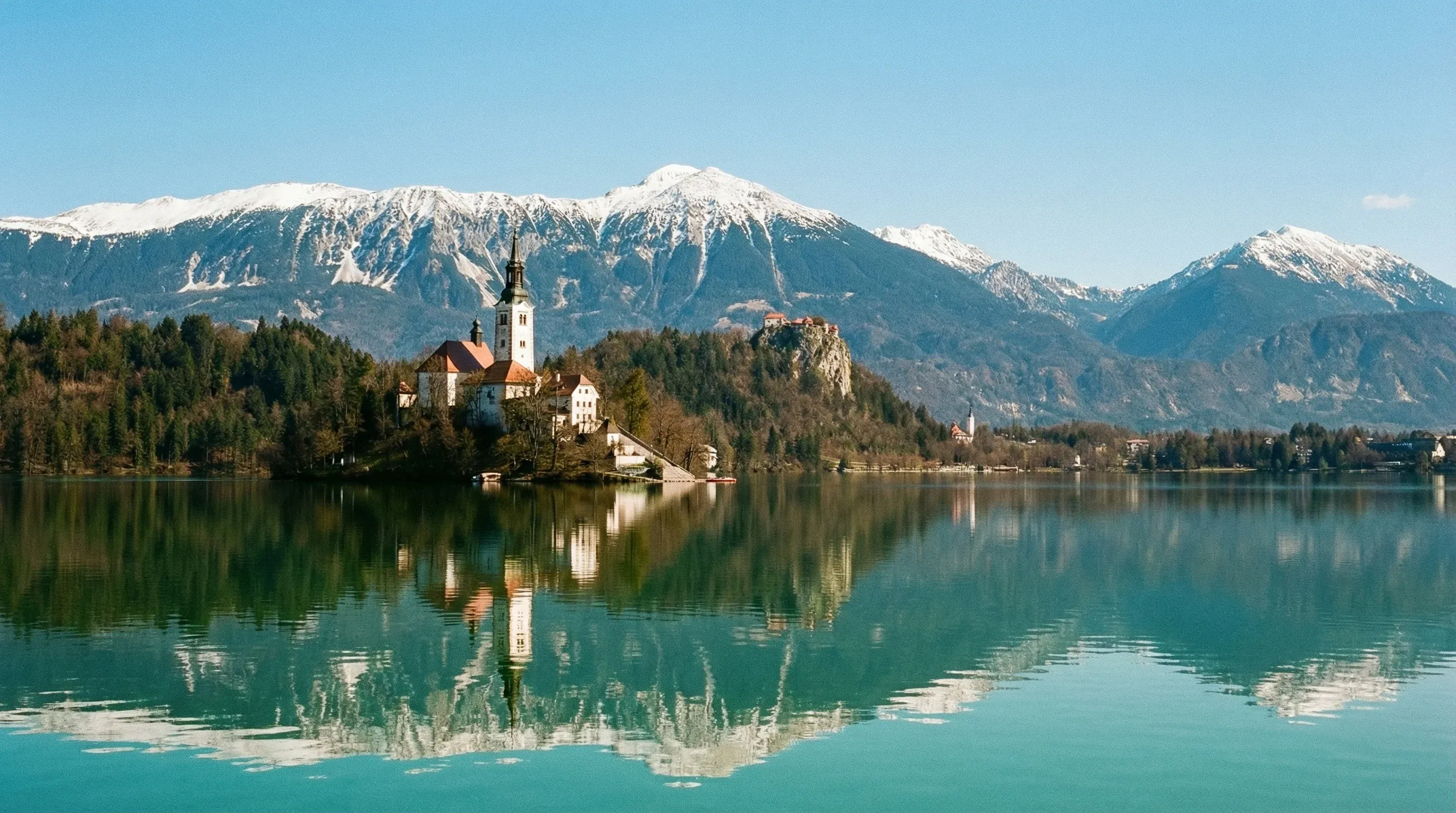 View of Lake Bled featuring the island church and the medieval castle on a cliff against the Julian Alps.