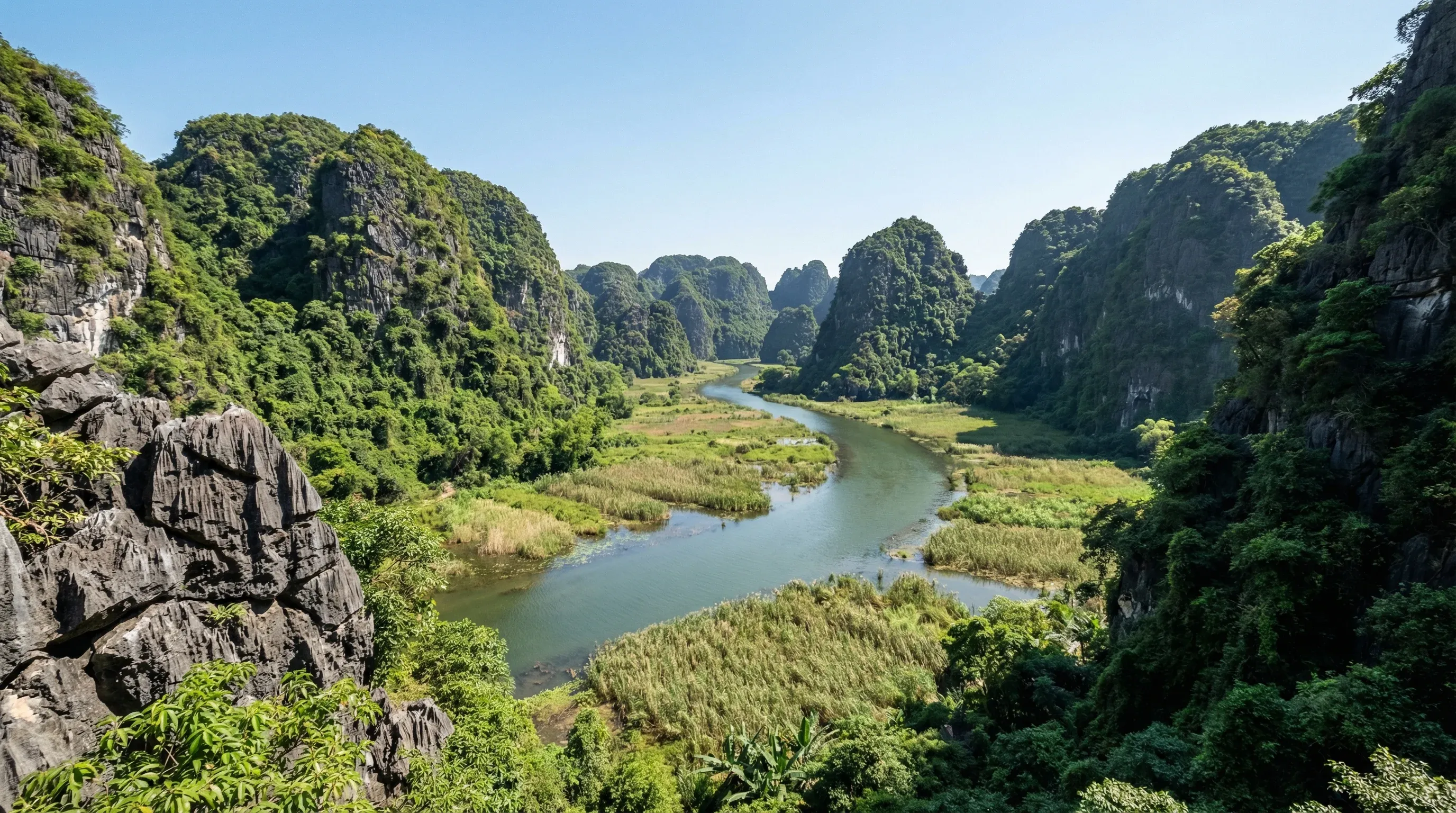 A winding river flowing through a valley of steep limestone karst mountains covered in green foliage in Ninh Binh, northern Vietnam.