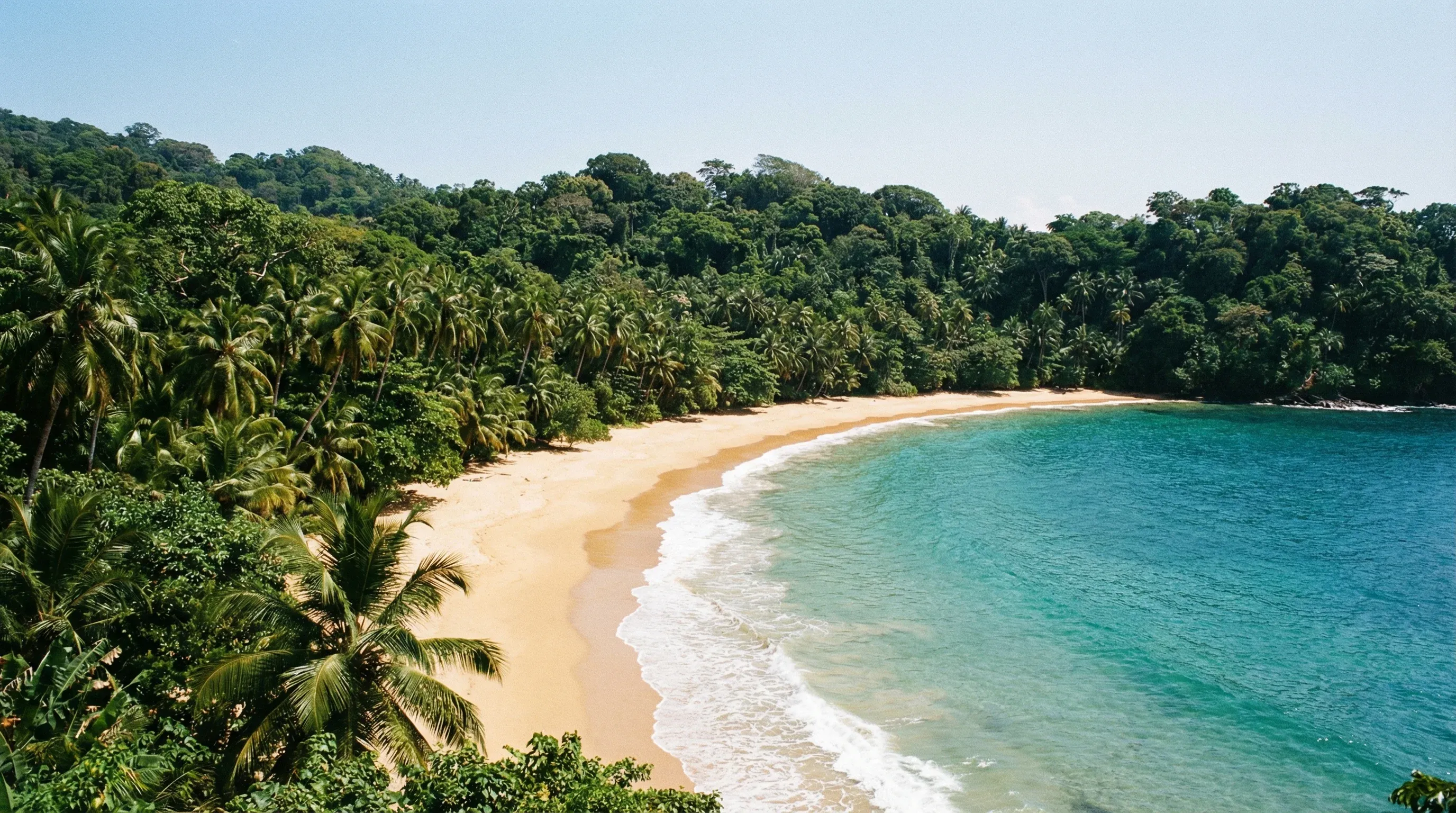 High-angle view of the crescent-shaped Praia Banana beach on Príncipe Island, surrounded by dense green jungle and turquoise ocean water.