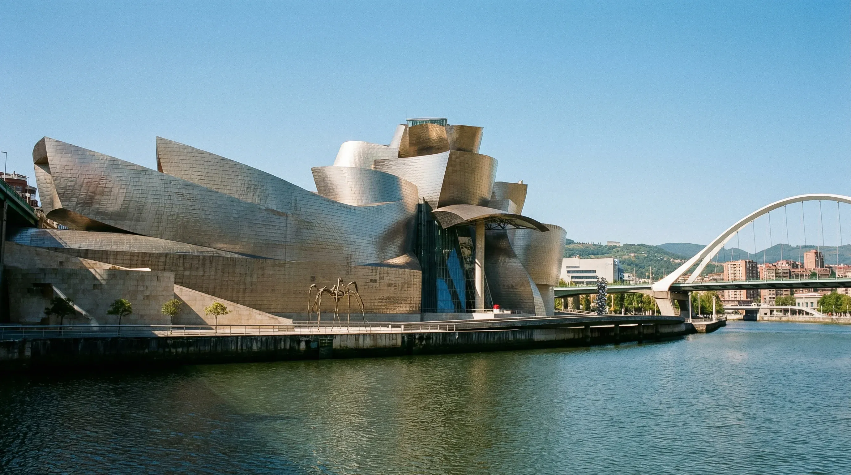 The titanium-clad Guggenheim Museum in Bilbao viewed from across the Nervion River under a bright blue sky.