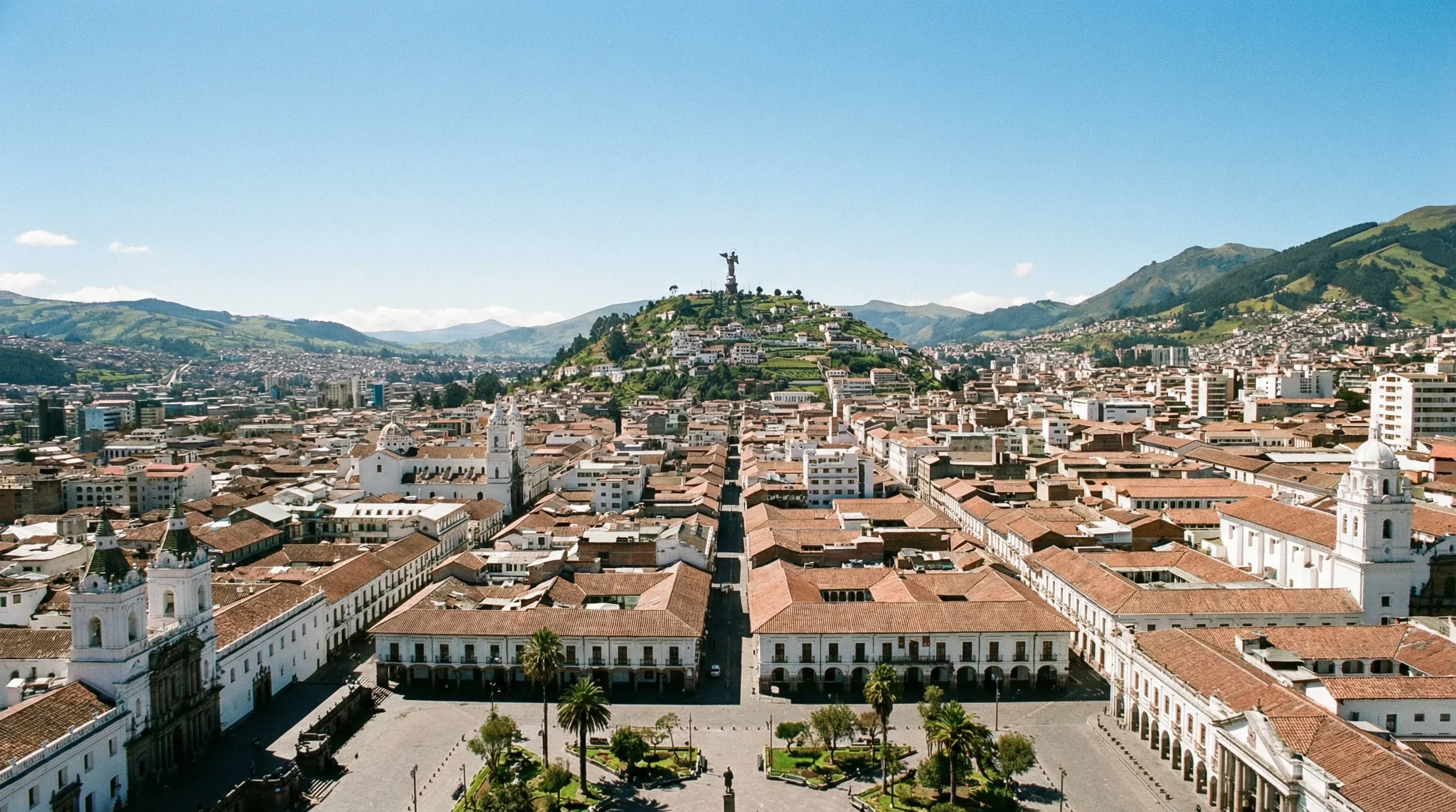 An expansive view of a colonial city with red roofs and white buildings, with a large winged statue on a hill in the distance.