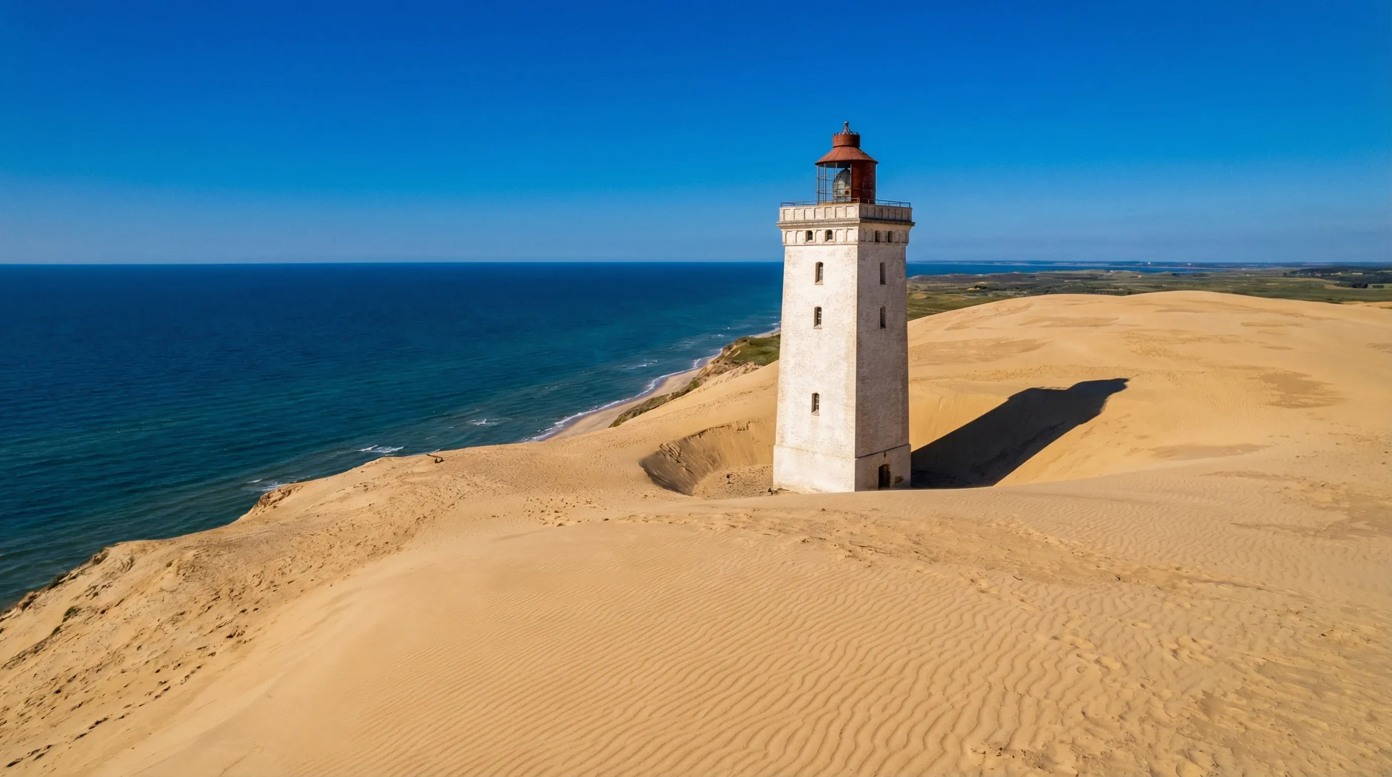 A solitary white lighthouse surrounded by high sand dunes on a cliff overlooking the blue North Sea.