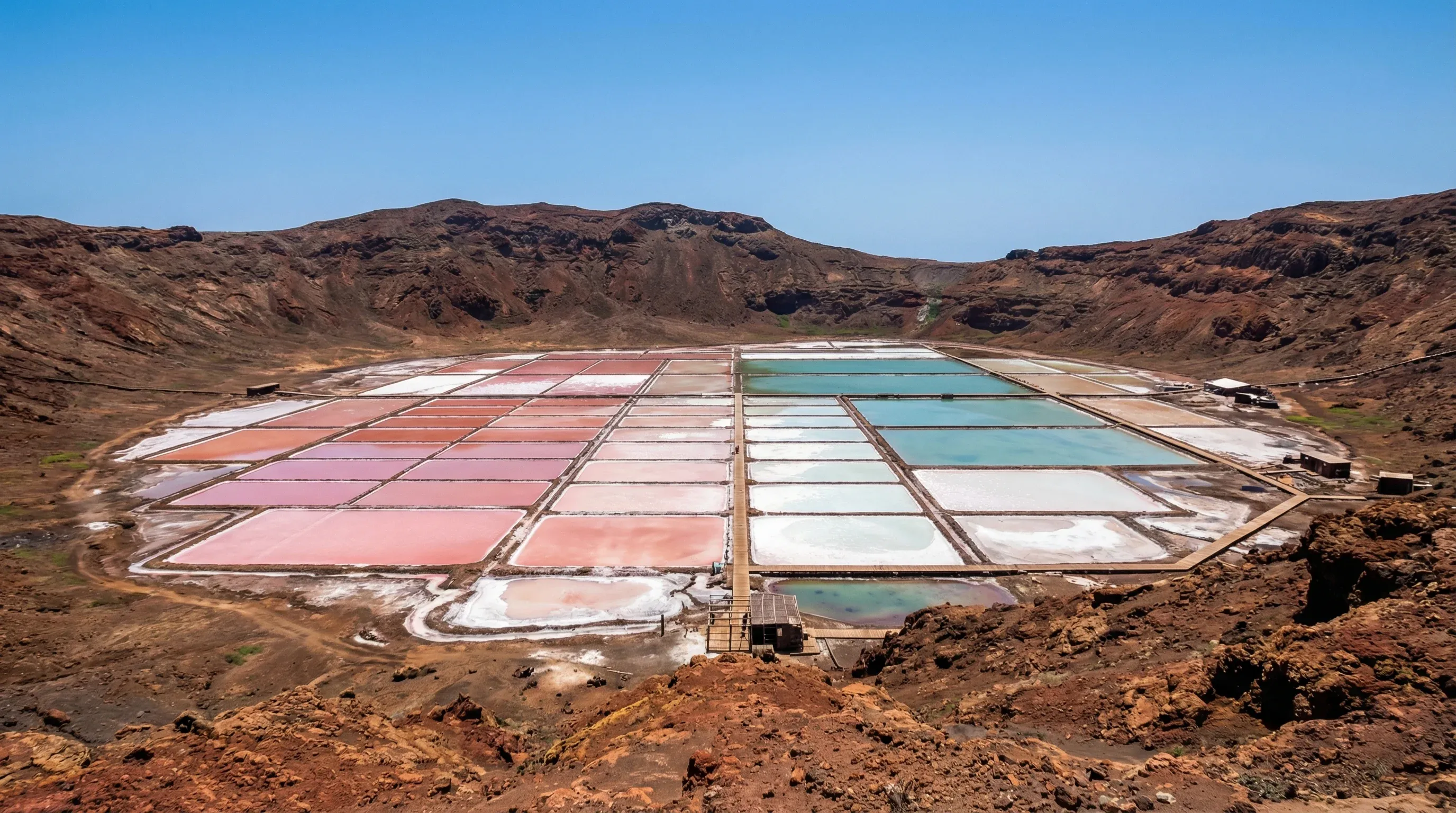 An elevated view of pink and white salt evaporation ponds inside a volcanic crater under a bright midday sun on Sal Island.