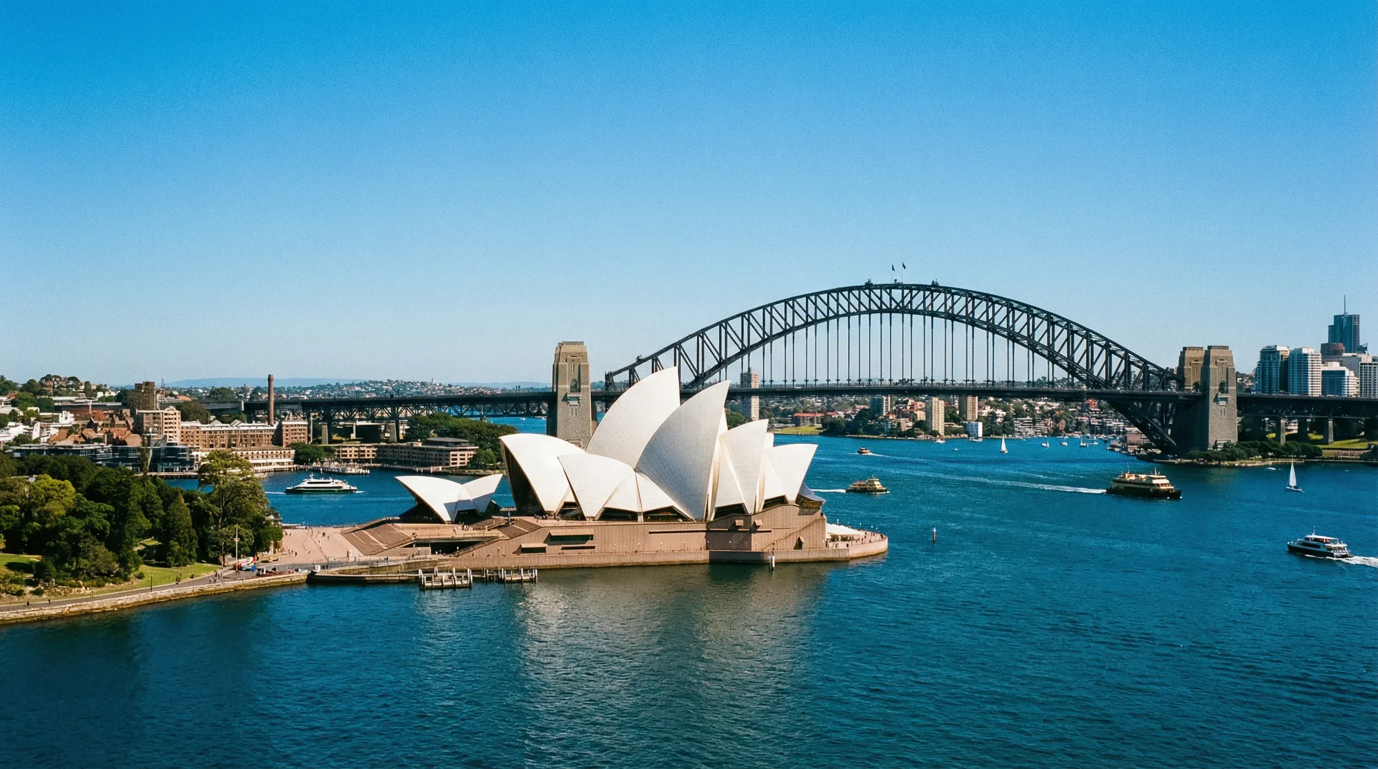 The white sails of the Sydney Opera House and the steel arch of the Sydney Harbour Bridge under a bright blue sky.