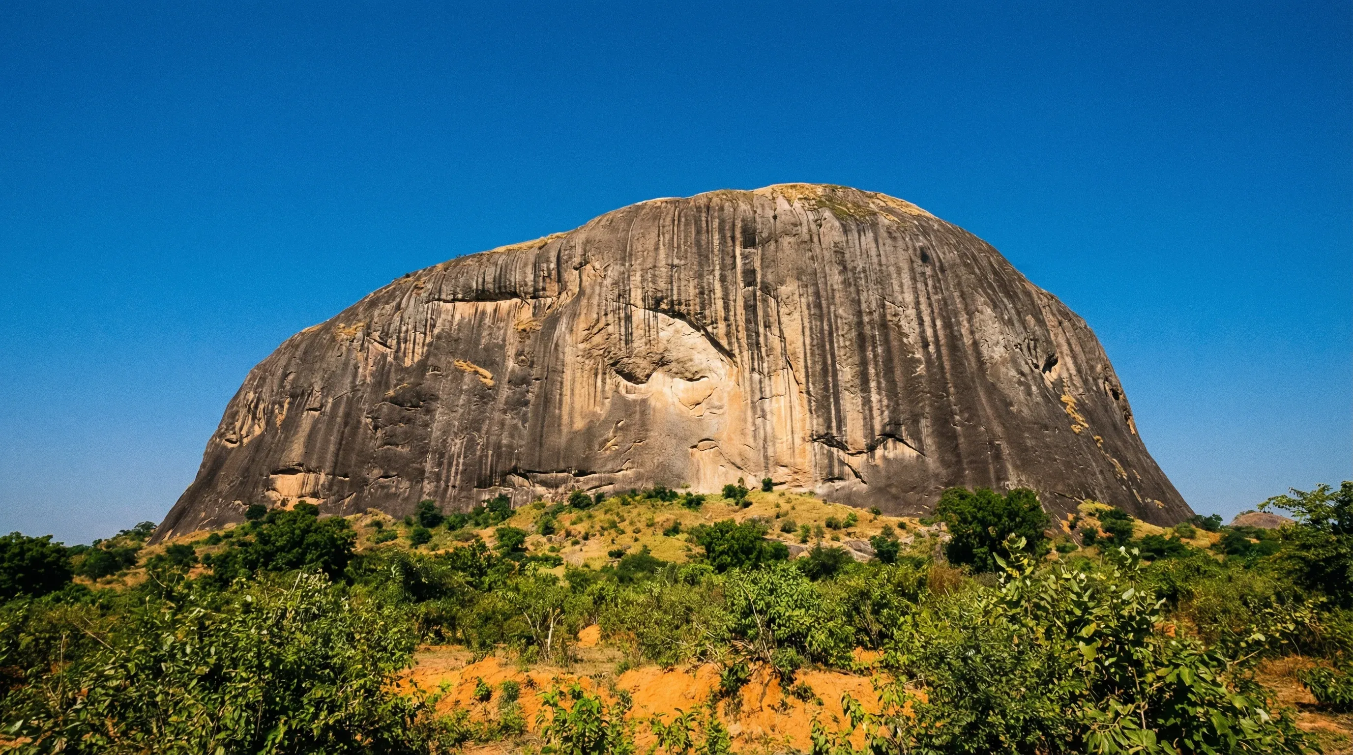 A massive granite monolith known as Zuma Rock rises from the green savanna under a clear blue sky in central Nigeria.