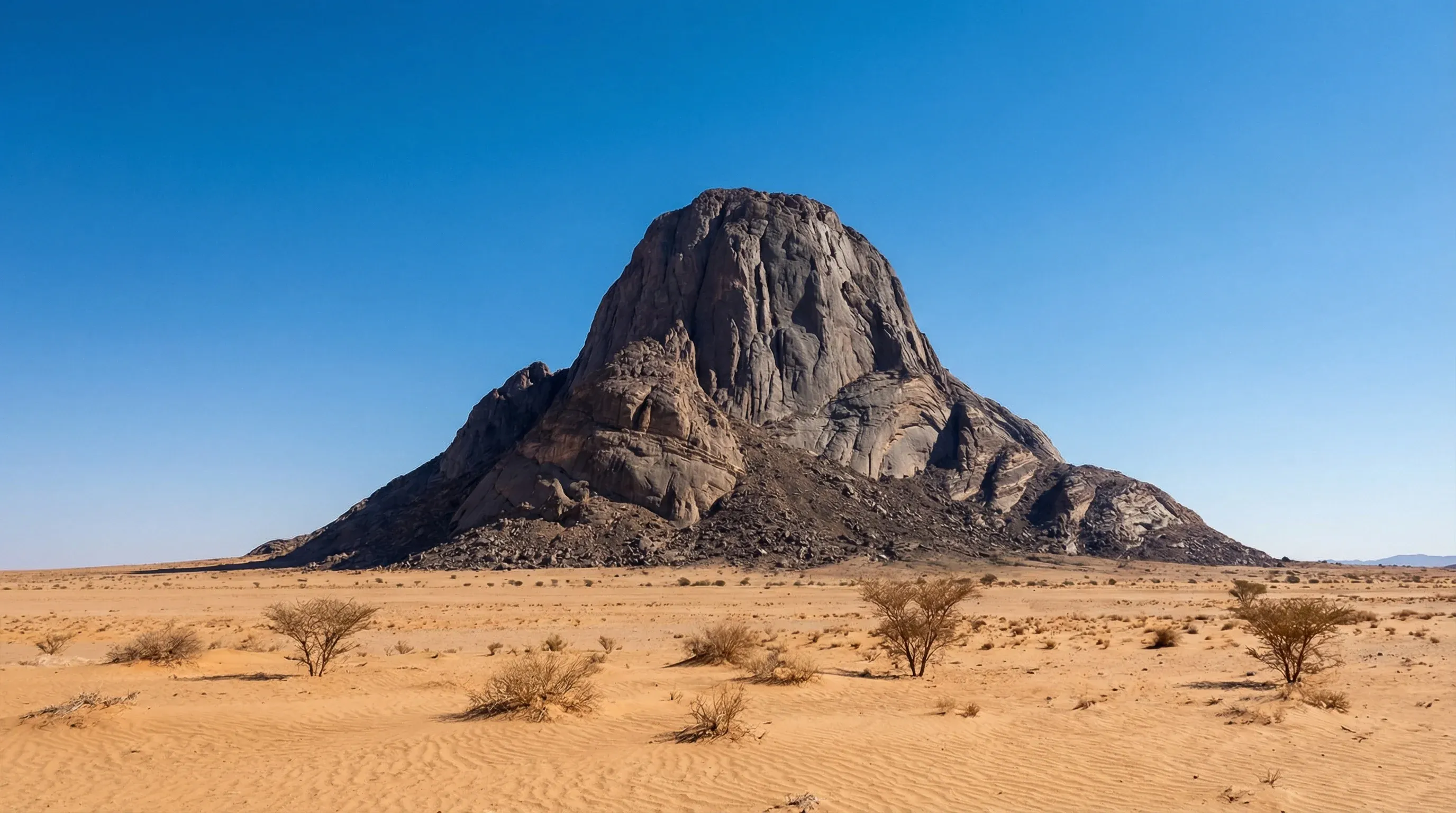 The massive dark granite monolith of Ben Amera rising from a flat, sandy desert plain under a clear blue sky in northern Mauritania.