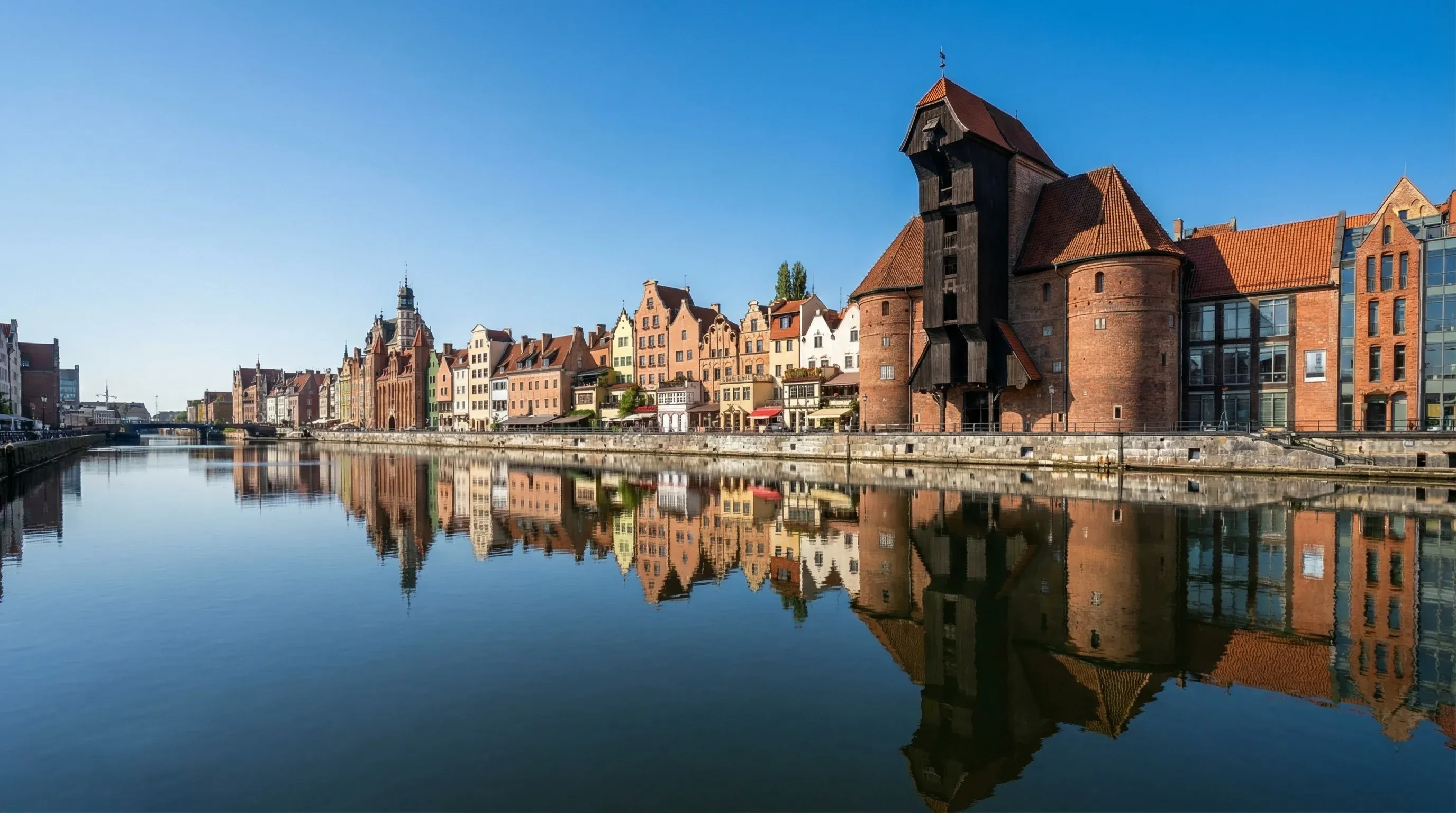 The historic 15th-century wooden crane and colorful narrow merchant houses along the Motława River in Gdańsk under a clear blue sky.