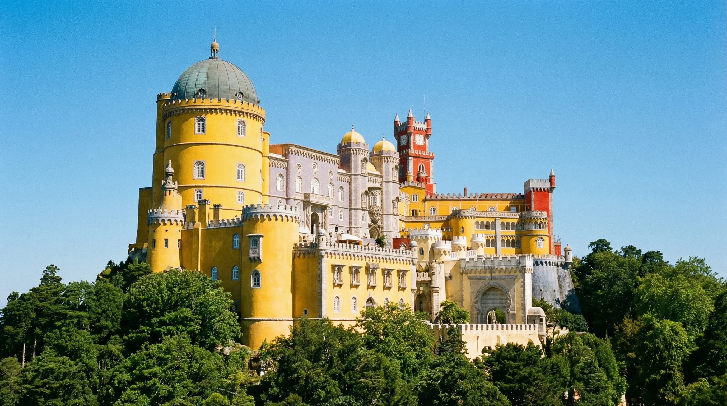 The colorful red and yellow towers of Pena Palace in Sintra, surrounded by a green forest under a blue sky.