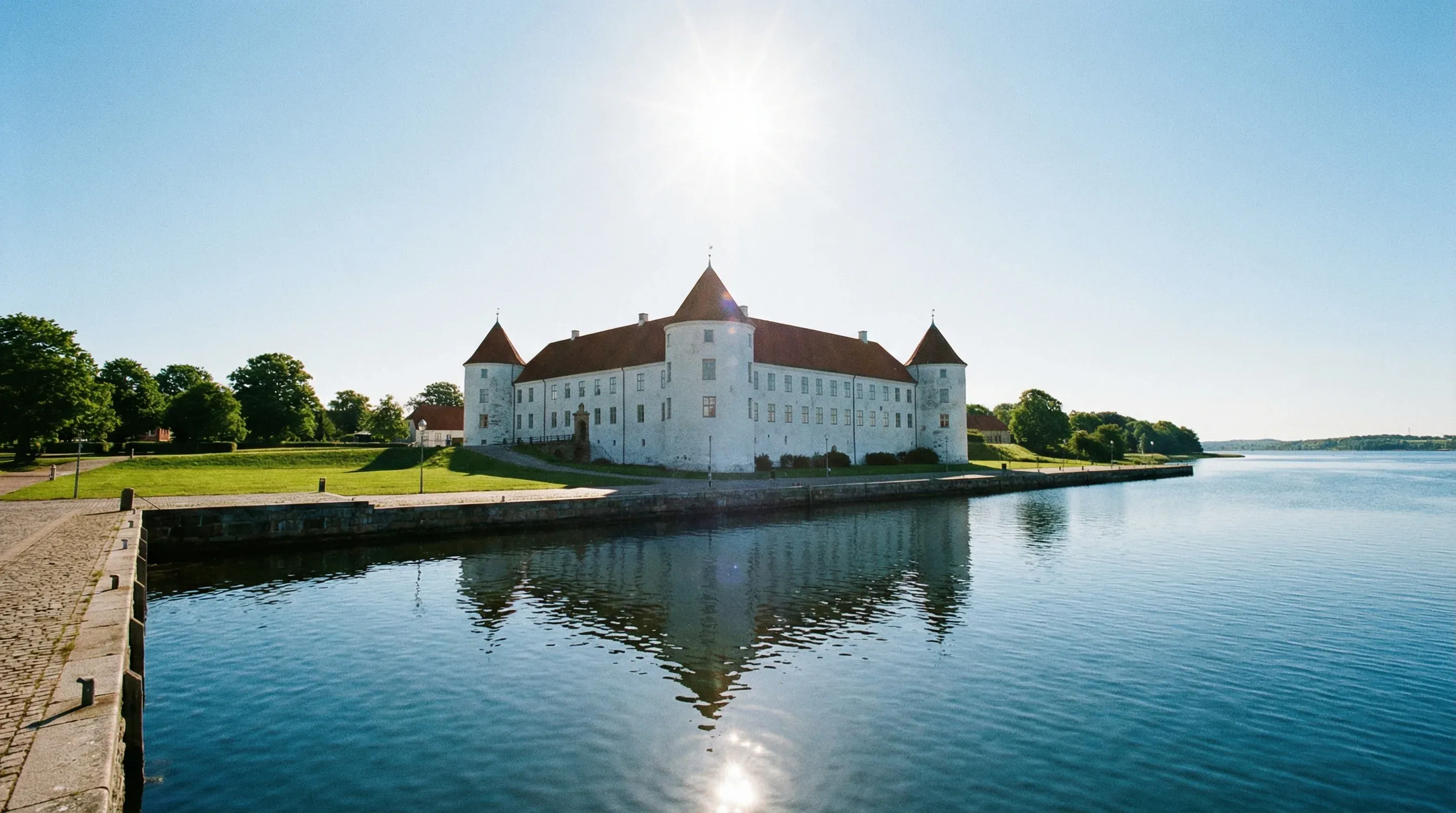 The white-walled Sønderborg Castle with red roofs located on the edge of a blue waterway under a clear sky.