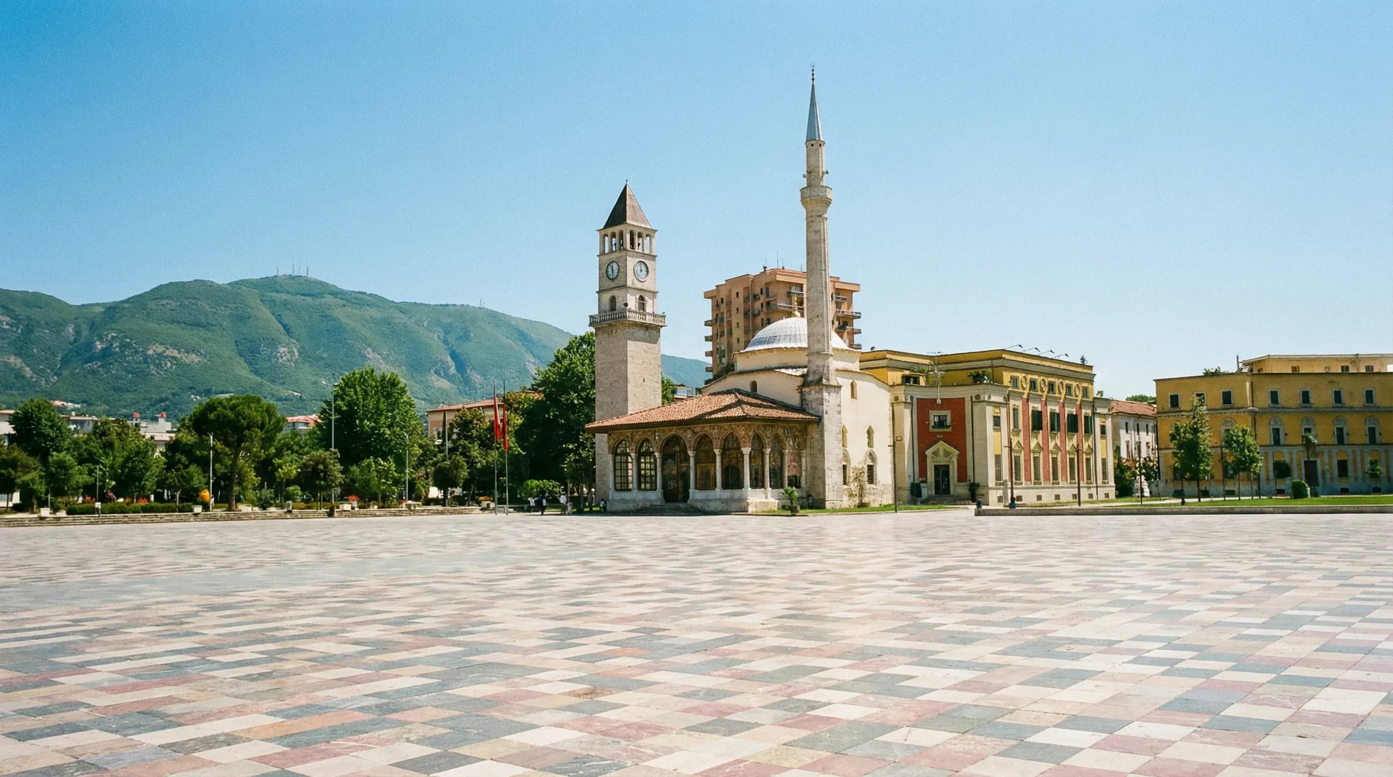 Skanderbeg Square in Tirana featuring the historic Et'hem Bey Mosque and the stone Clock Tower against a clear blue sky.