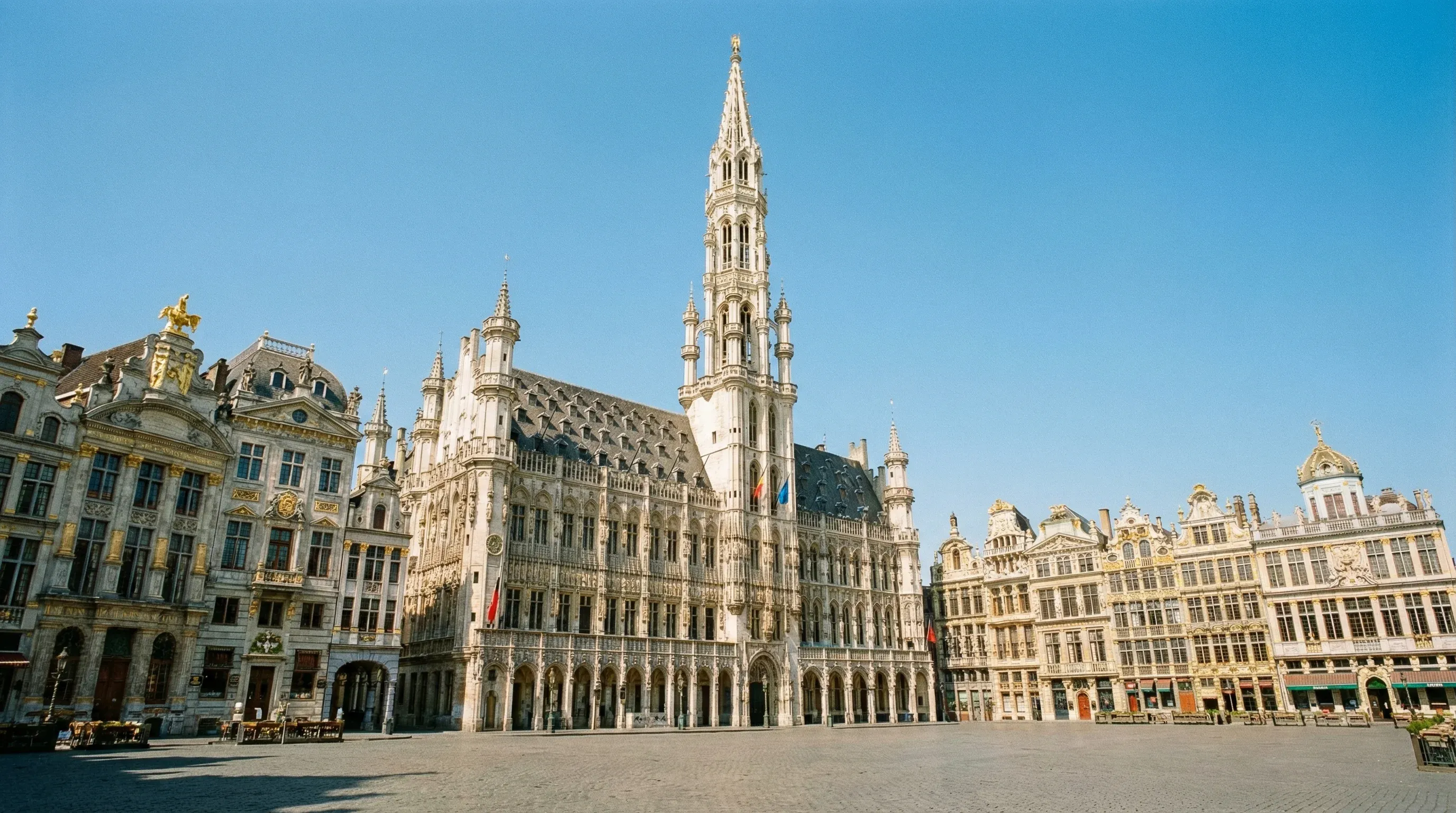 A wide-angle view of the Grand Place in Brussels, showcasing the Gothic Town Hall and ornate guildhouses under a clear blue sky.