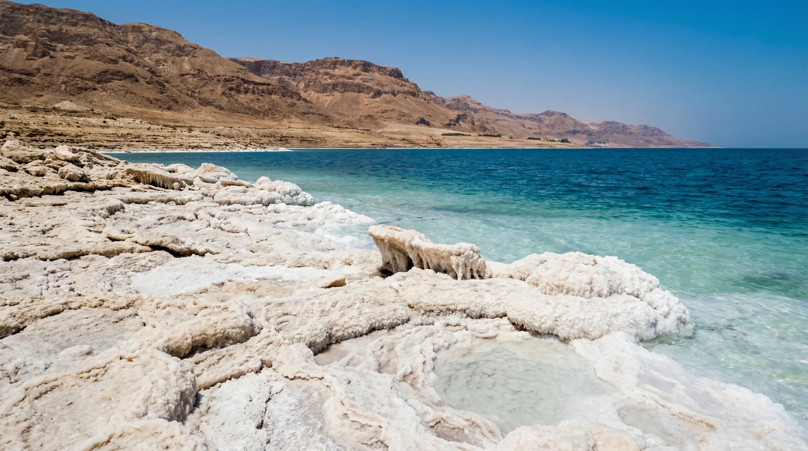 White salt formations crust the shoreline of the deep blue Dead Sea, with the hazy mountains of the Rift Valley in the background.