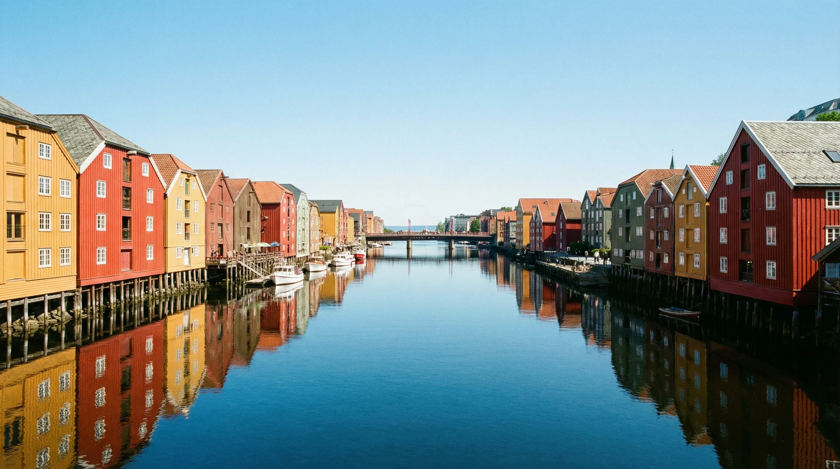 A row of colorful red and yellow wooden buildings on stilts reflected in the calm Nidelva river in Trondheim.