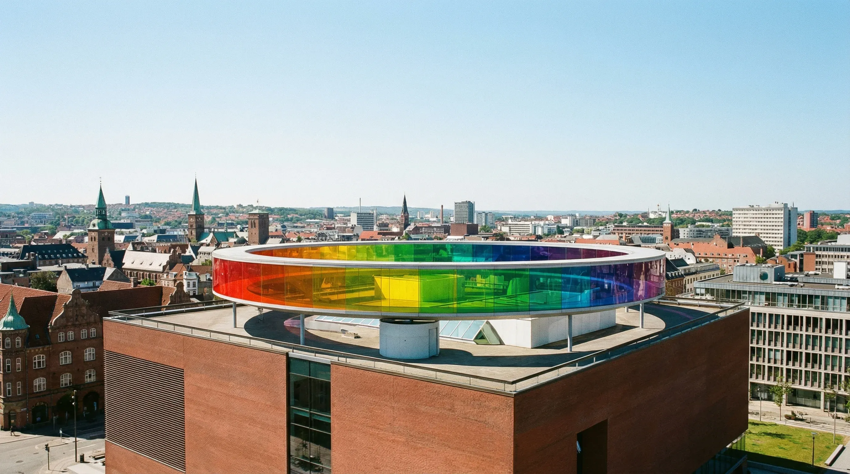 The circular rainbow-colored glass walkway on top of the ARoS Aarhus Art Museum building under a clear sky.
