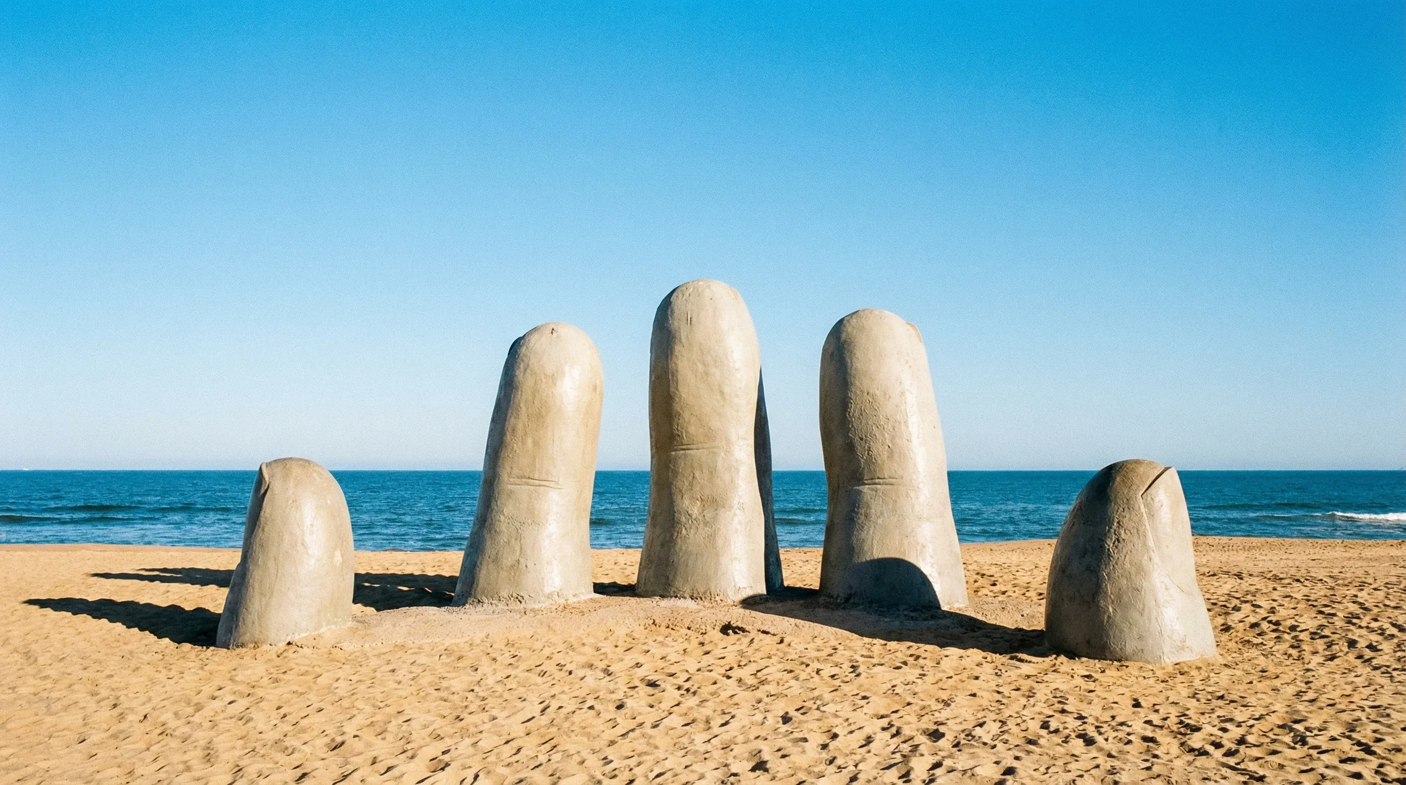 A large concrete sculpture of a hand emerging from the sand on a sunlit beach with the blue ocean in the distance.