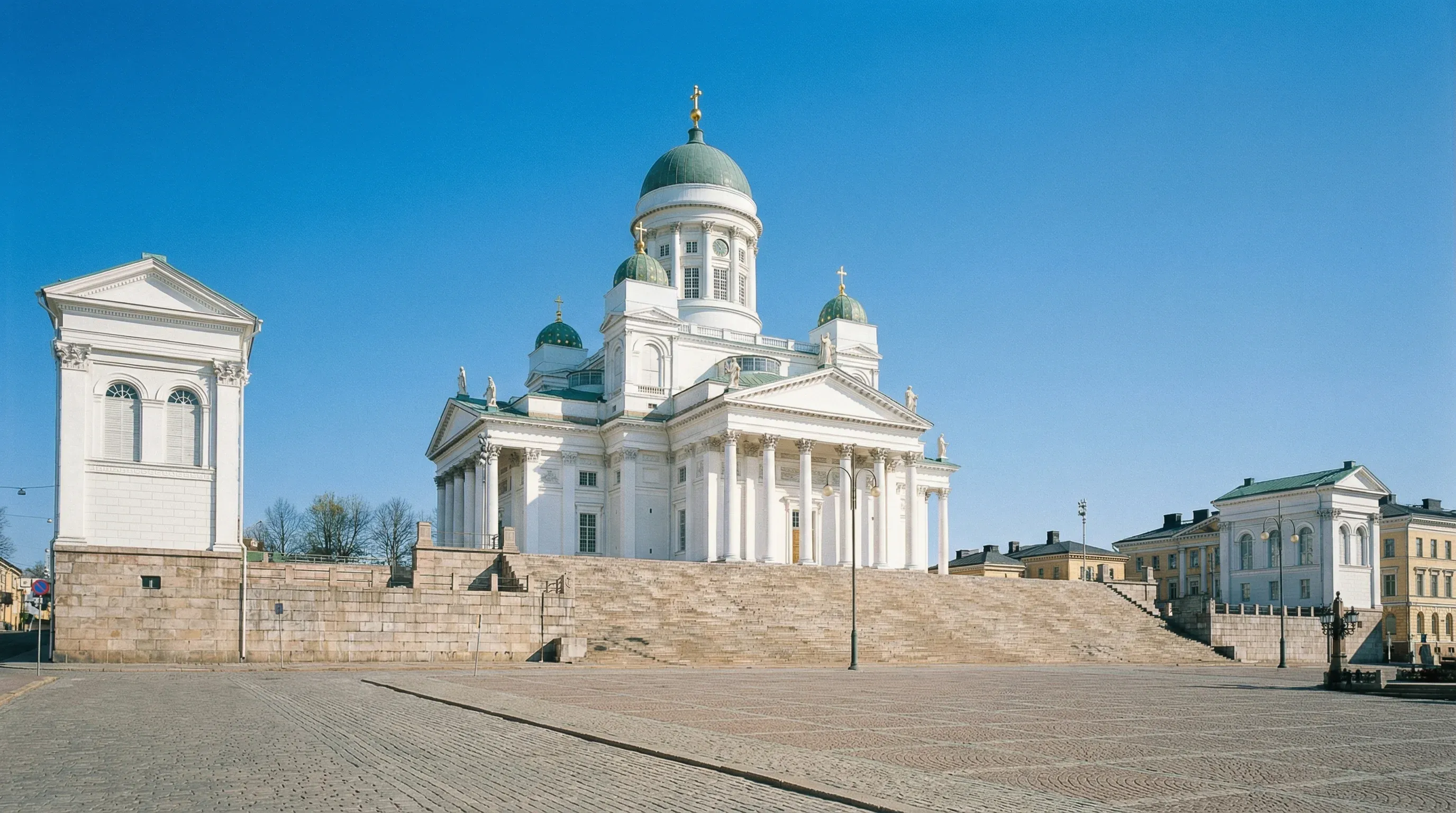 A large white neoclassical cathedral with green domes and gold accents stands prominently at the top of a stone staircase.