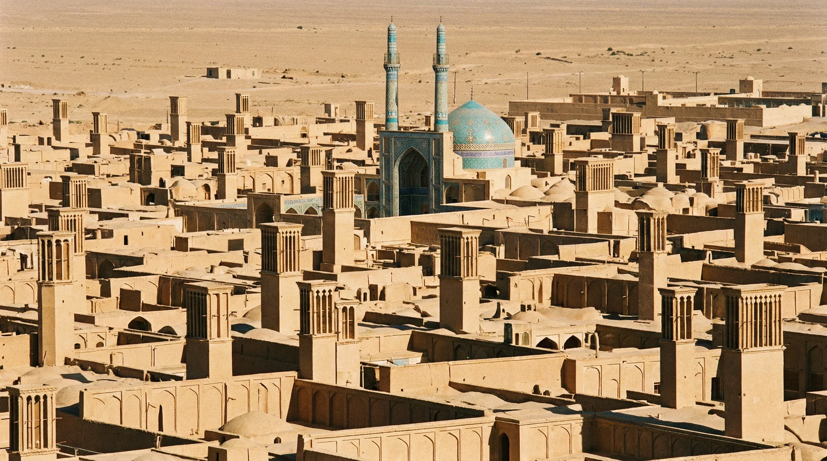 A view across the mud-brick rooftops of Yazd featuring traditional windcatchers and the tall blue minarets of the Jameh Mosque.